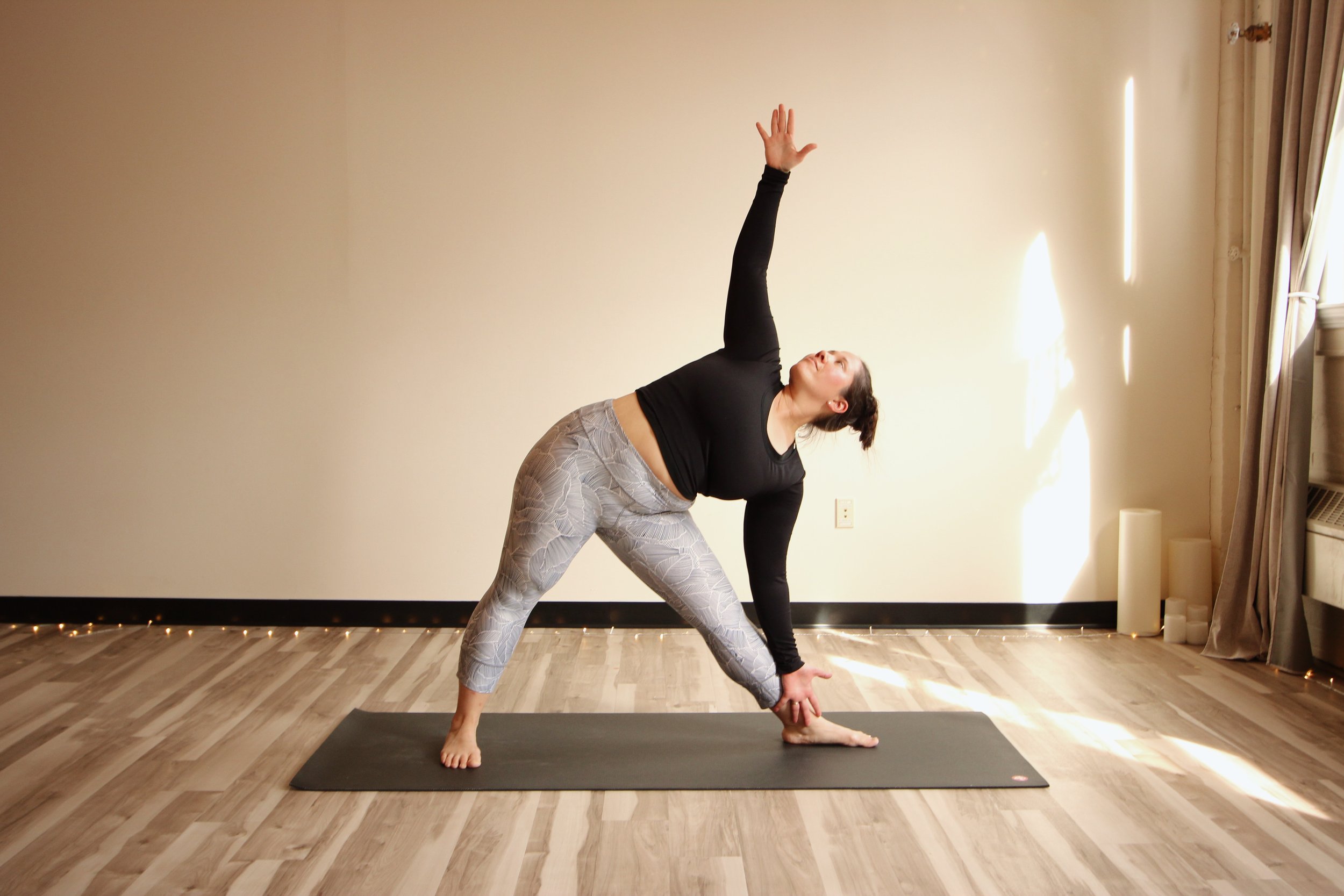 A woman practicing yoga indoors on a black yoga mat. She is in a triangle pose, with one hand reaching upward and the other touching her foot, on a wooden floor against a blank wall with sunlight coming through a curtain on the right.