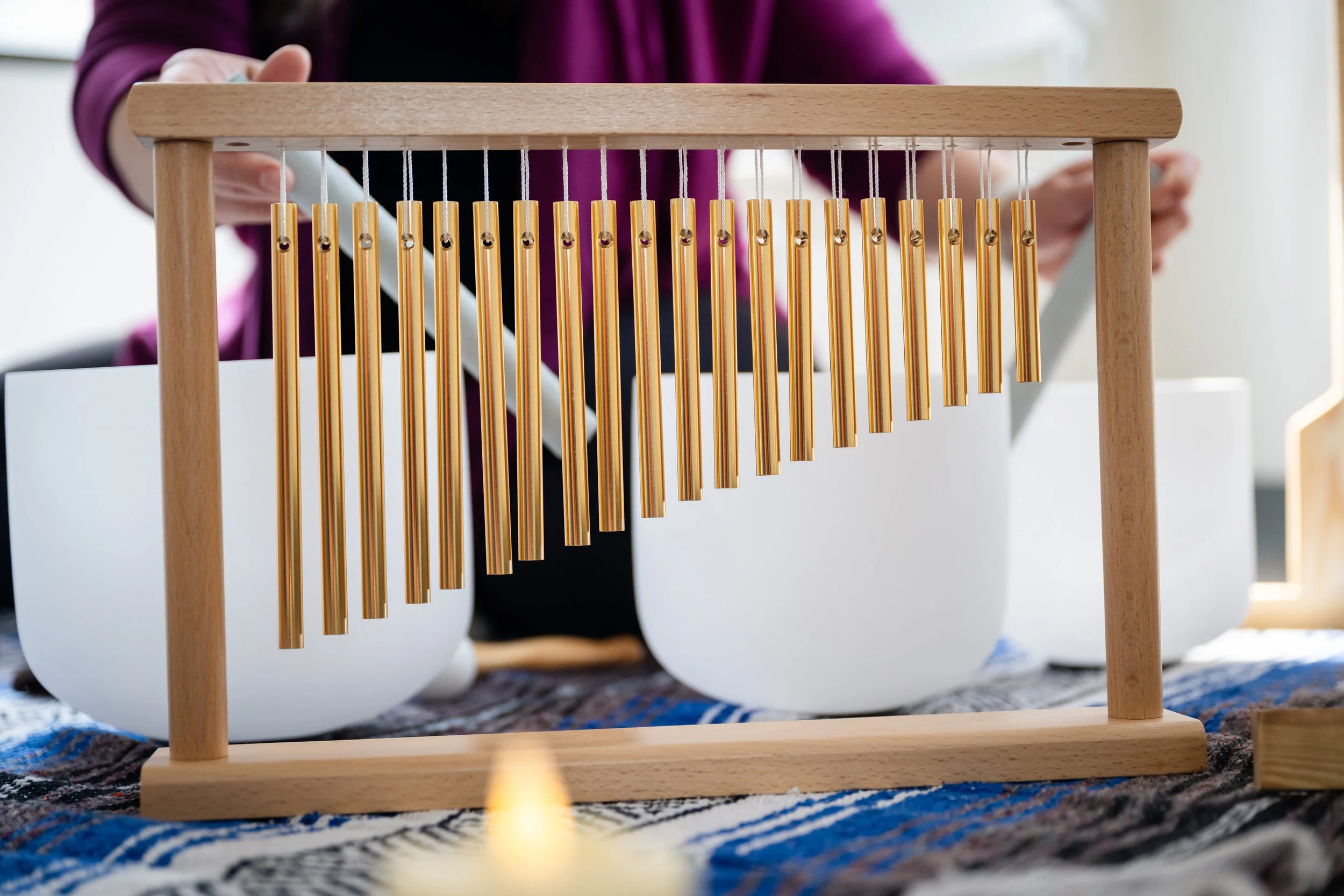 Close-up of a wooden frame with metallic chimes hanging from it, with a person in the background wearing a purple jacket.