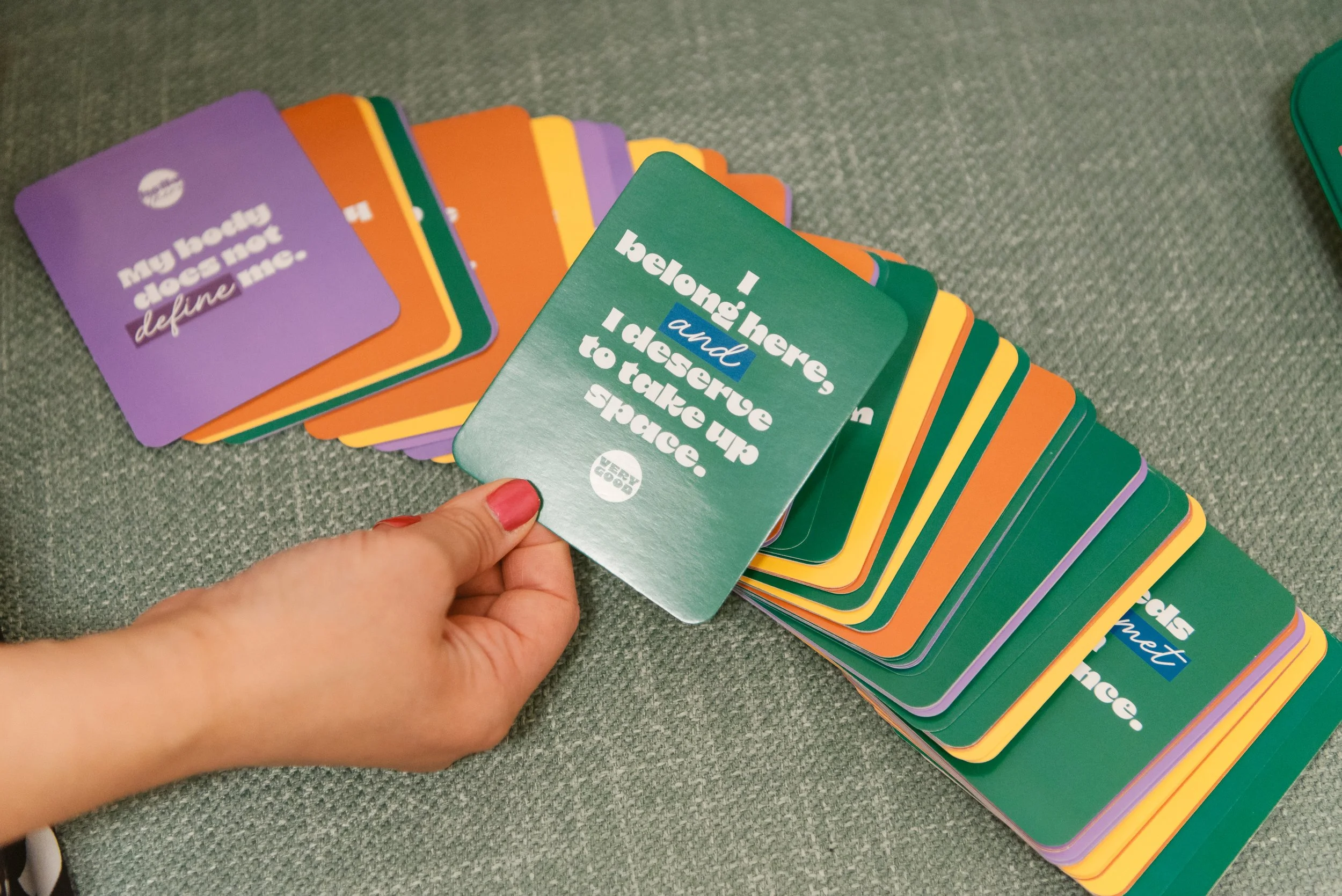 Person with red painted nails holding a green card from a colorful deck with the text 'Belonging, and I deserve up to take up to space.' The deck contains various other cards with different motivational messages, spread out on a gray surface.