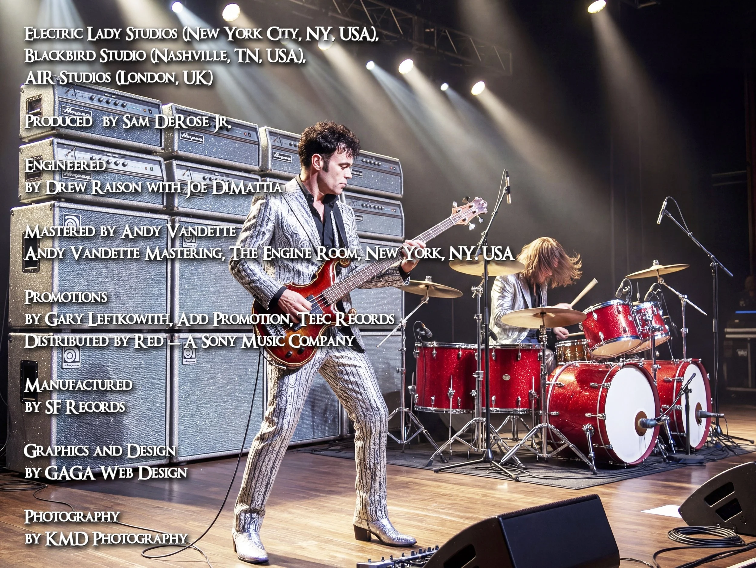 A musician with dark, curly hair, wearing a silver suit with black and white patterns, playing a red electric guitar on stage. Behind him are large amplifier stacks, and other musicians are playing drums. Stage lights are shining down, and the settin