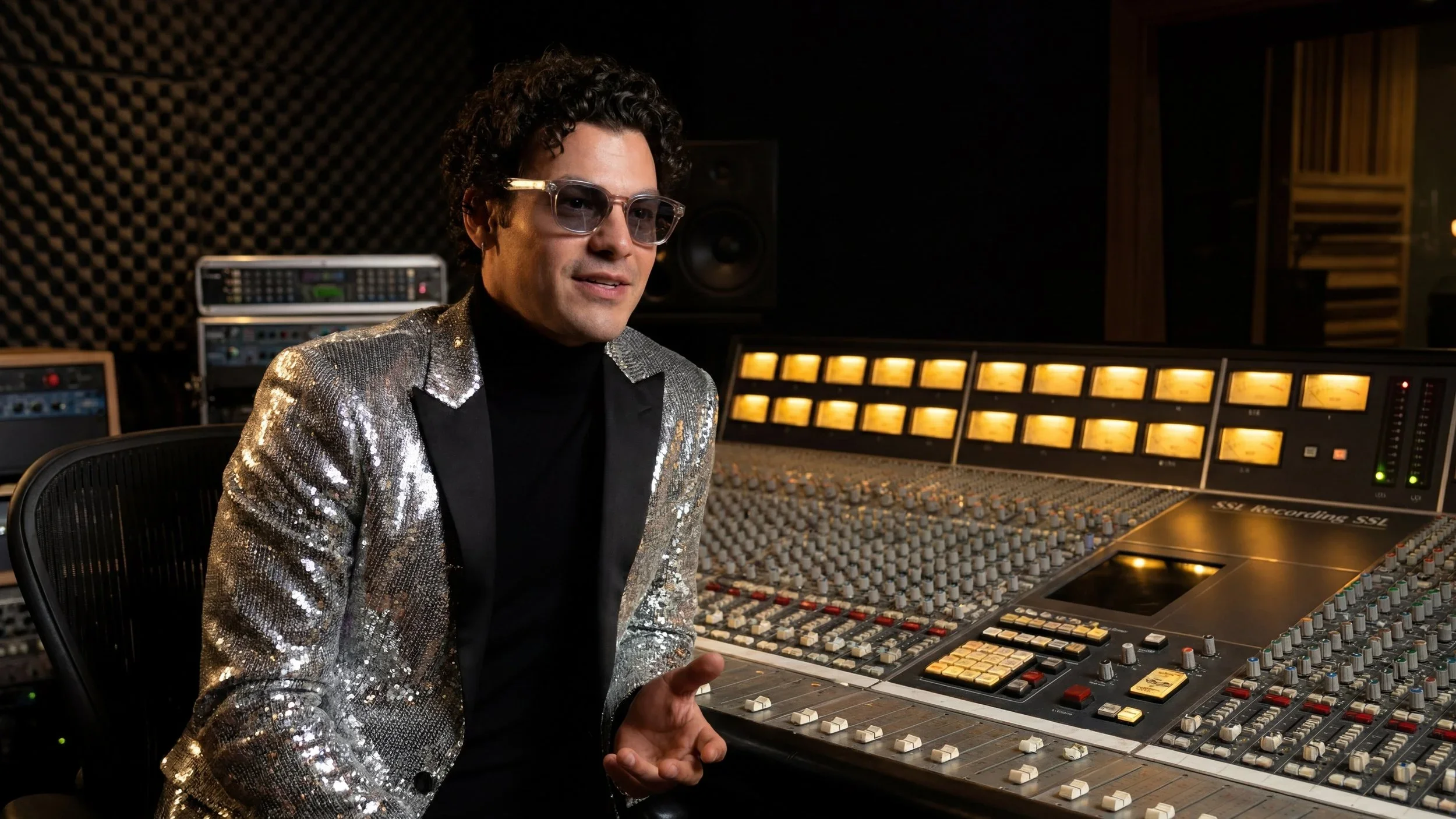 A man wearing sunglasses and a silver sequin blazer sitting in a recording studio, surrounded by an audio mixing console and sound equipment.