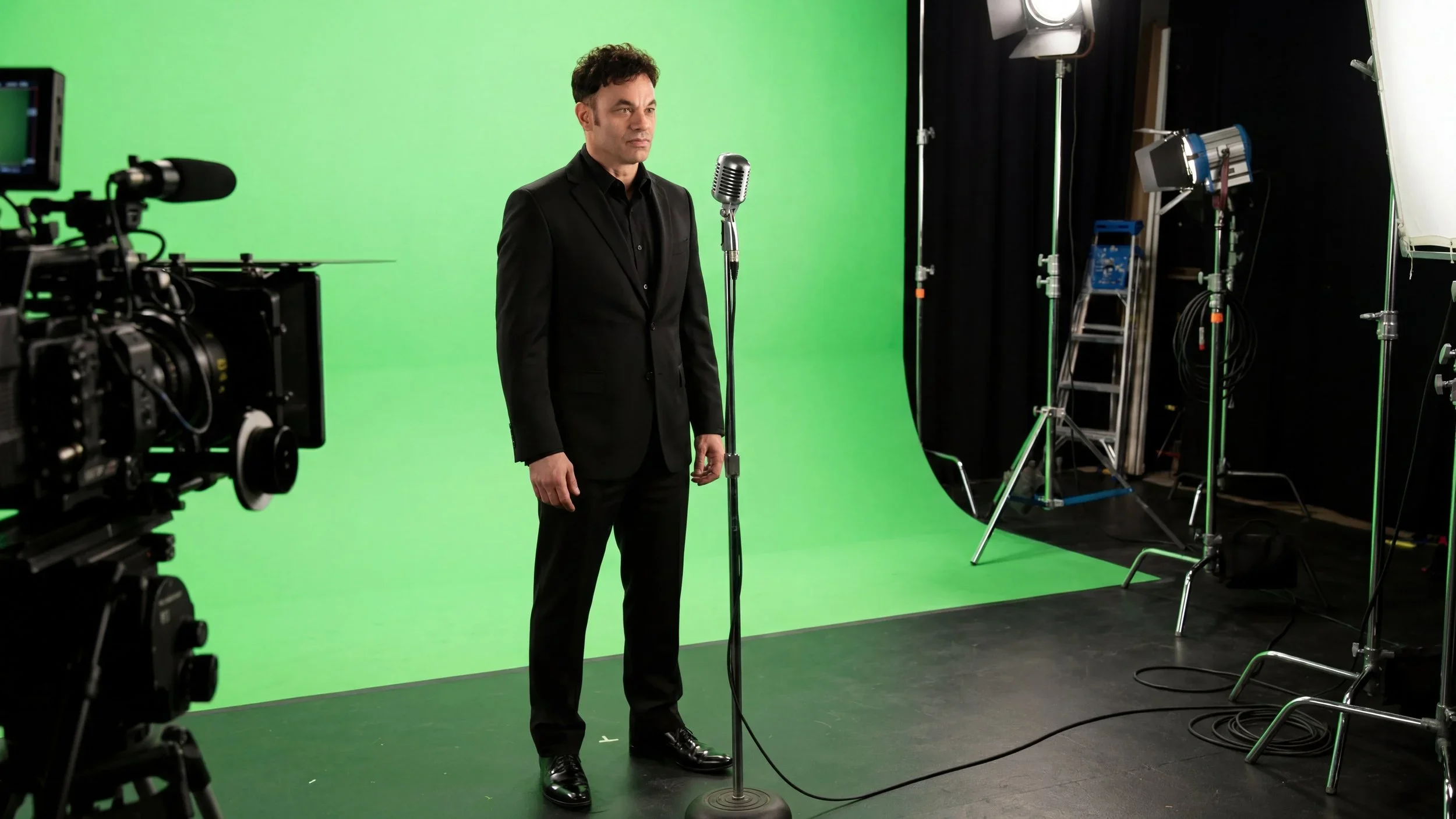 Man in black suit standing in front of green screen with vintage microphone, surrounded by film studio lighting and equipment.