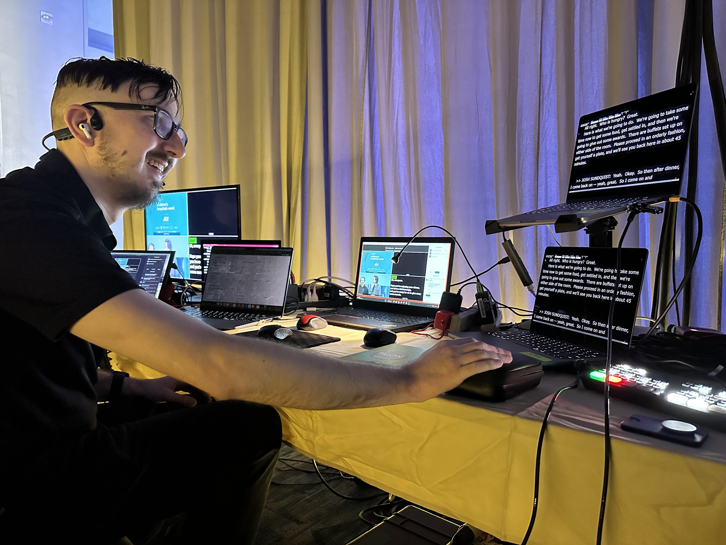 A captioning technician monitors live captions on several laptops while smiling. He is wearing two headsets and has several monitors and laptops on the table in front of him.