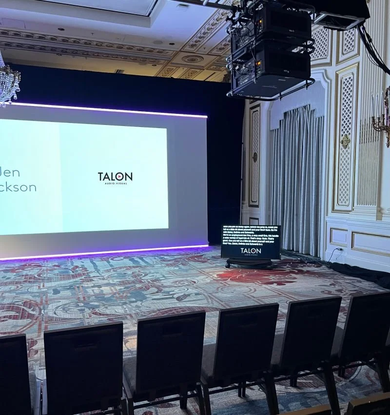 A large screen shows the Talon AV Logo, with a TV on a floor stand sitting below the large projector screen shows live captions of the event, with a Talon AV Logo on the screen. Projectors are rigged to the ceiling.