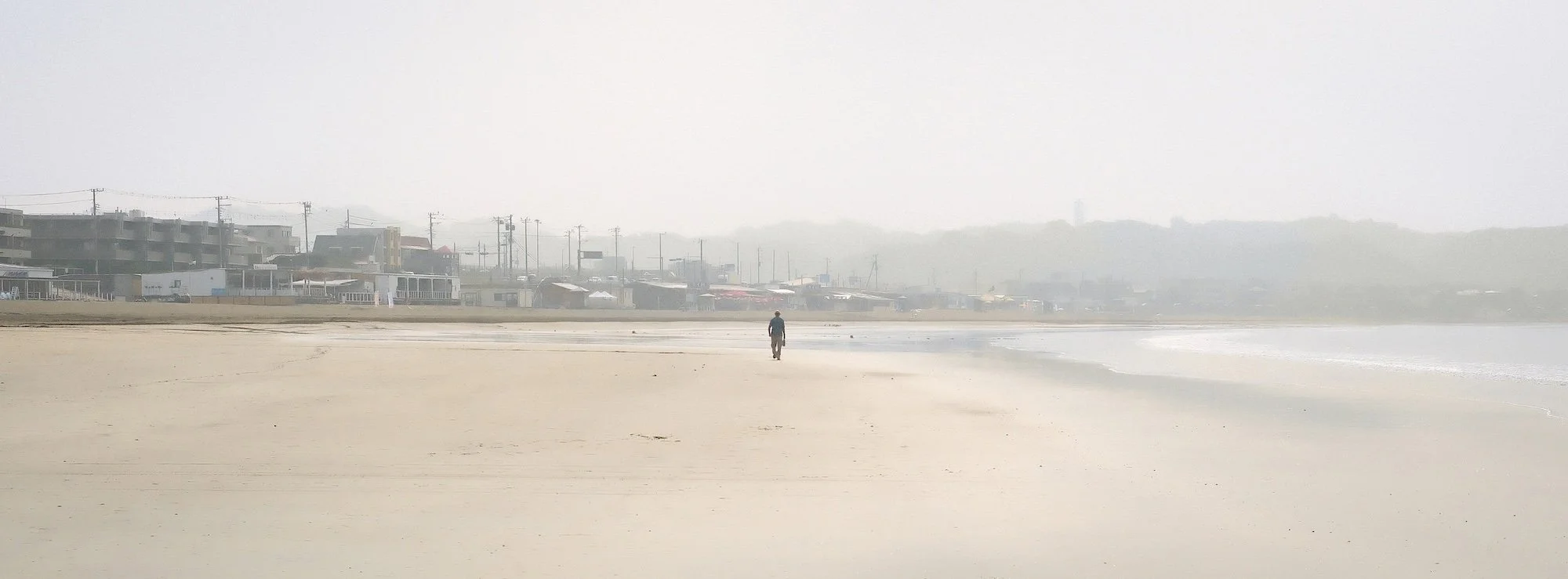 A person walking alone on the sandy beach with buildings and structures faintly visible through fog in the background.