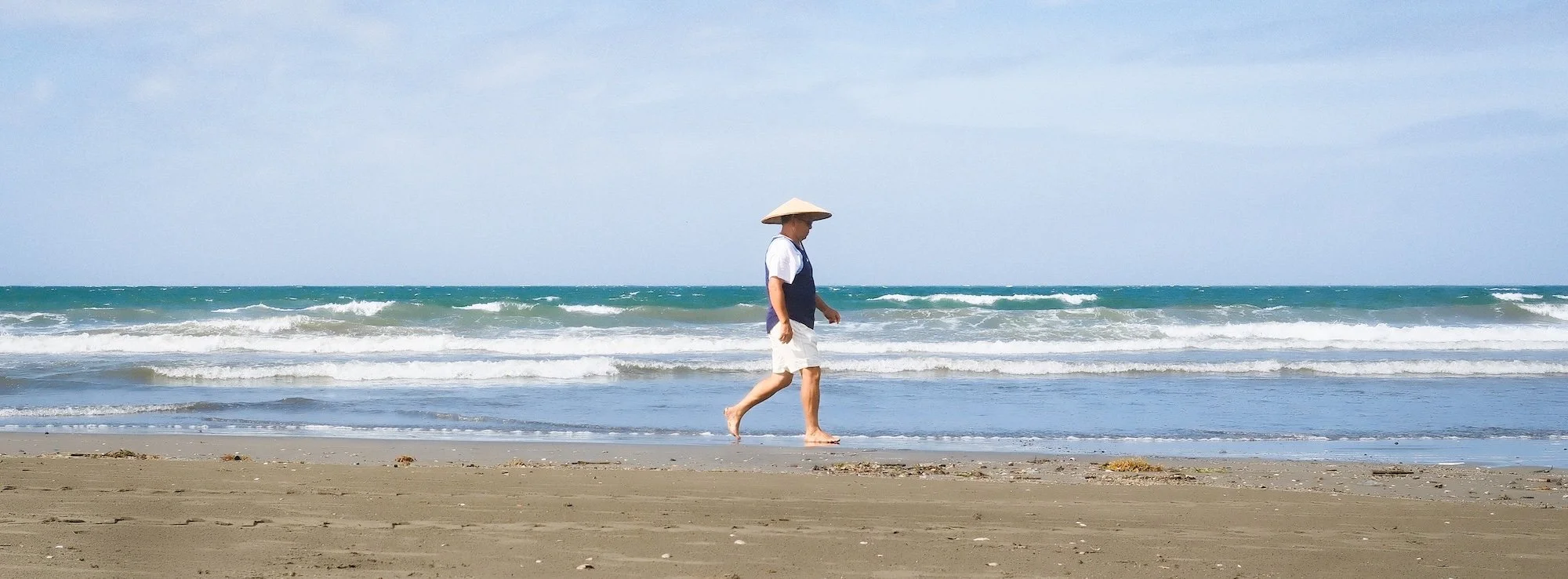 A person wearing a straw hat, white shorts, and a navy vest walking along the sandy beach near the shoreline with waves in the background.