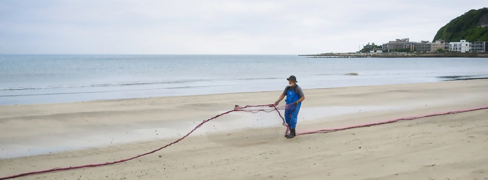 An elderly man with a gray beard wearing a black hat, gray shirt, and blue waterproof overalls stands on a sandy beach, holding a red fishing net. The ocean with gentle waves and a cloudy sky are in the background, along with some buildings on a hill