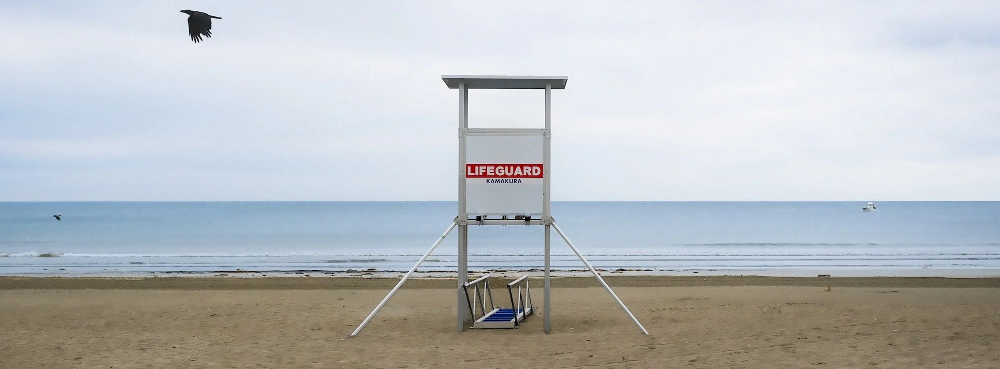 Empty lifeguard tower on a sandy beach with the ocean and a few boats in the background, overcast sky.