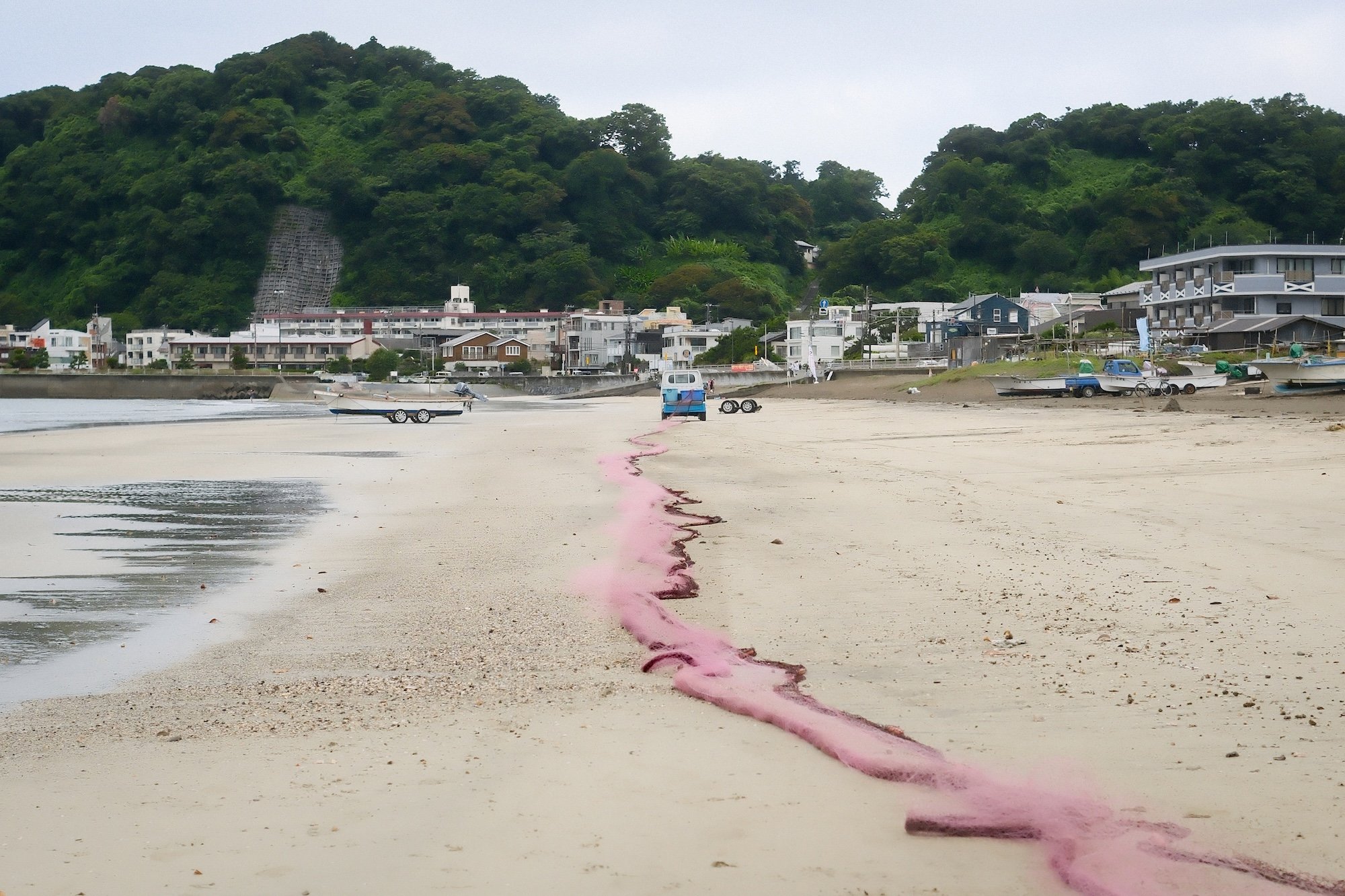 Pink oil spill on a sandy beach with boats and buildings in the background, hills, and overcast sky.