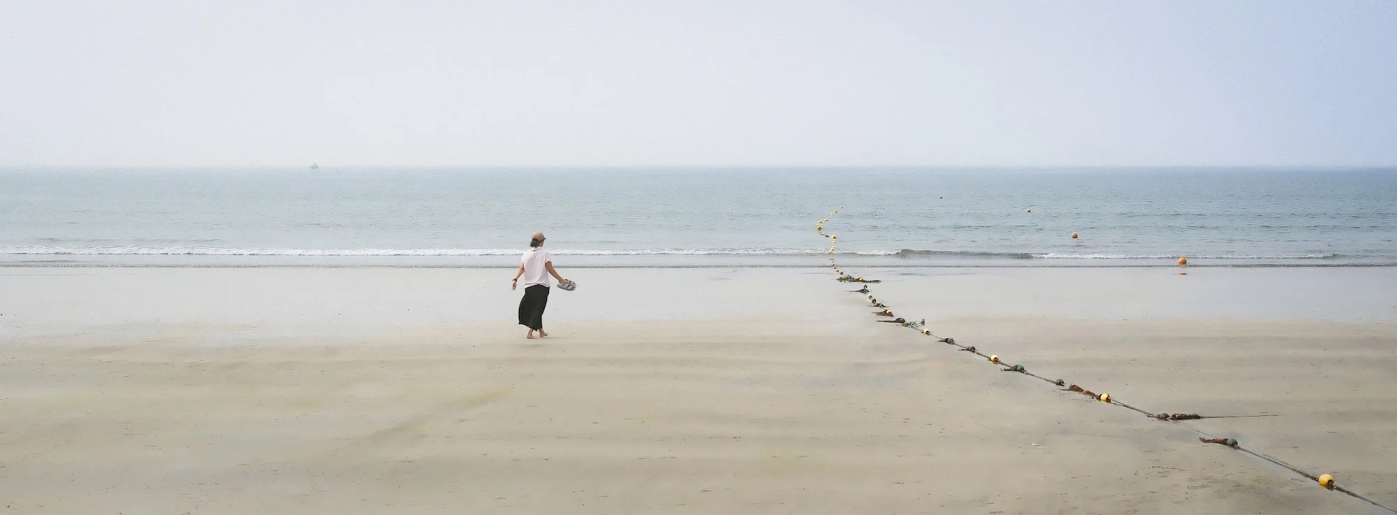 A person holding shoes, walking on a beach with a rope barrier extending into the water and the horizon in the distance.