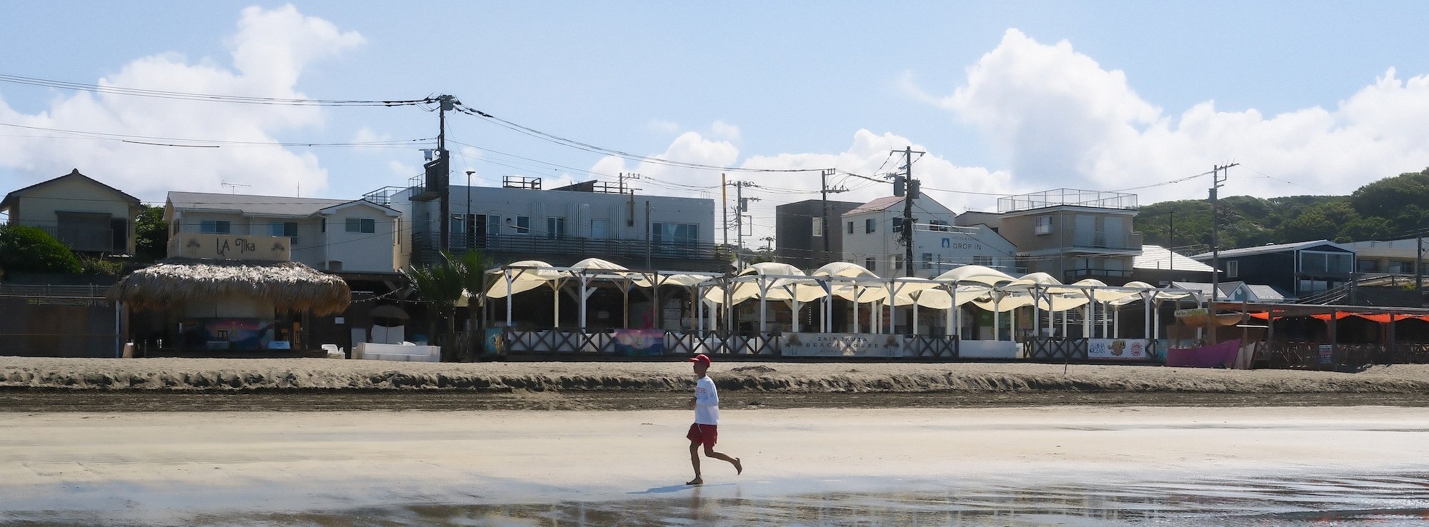Person running on wet sandy beach with beach houses, umbrellas, and a thatched hut along the shoreline under a blue sky with clouds.