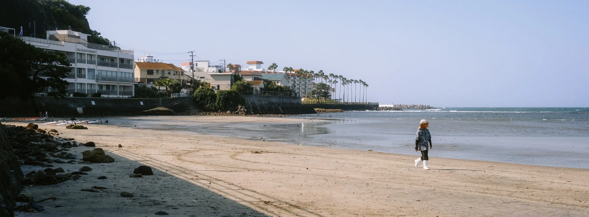 A person walking along a sandy beach near the ocean with residential buildings and palm trees in the background.