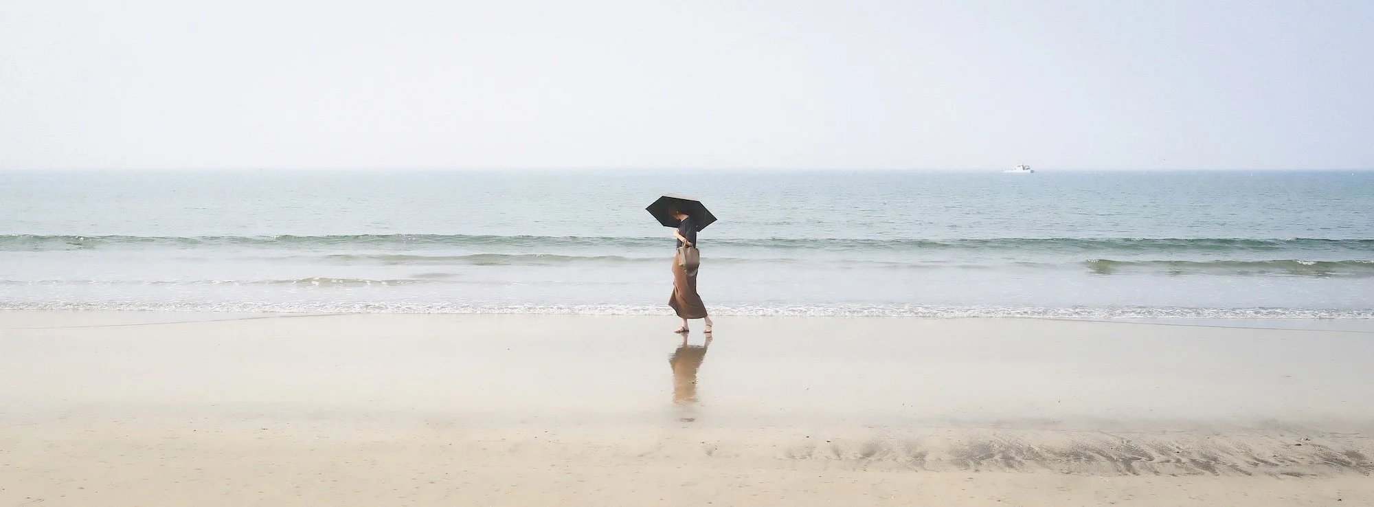 A person holding a black umbrella walking on a sandy beach near the ocean with small waves, overcast sky, and a distant boat on the water.