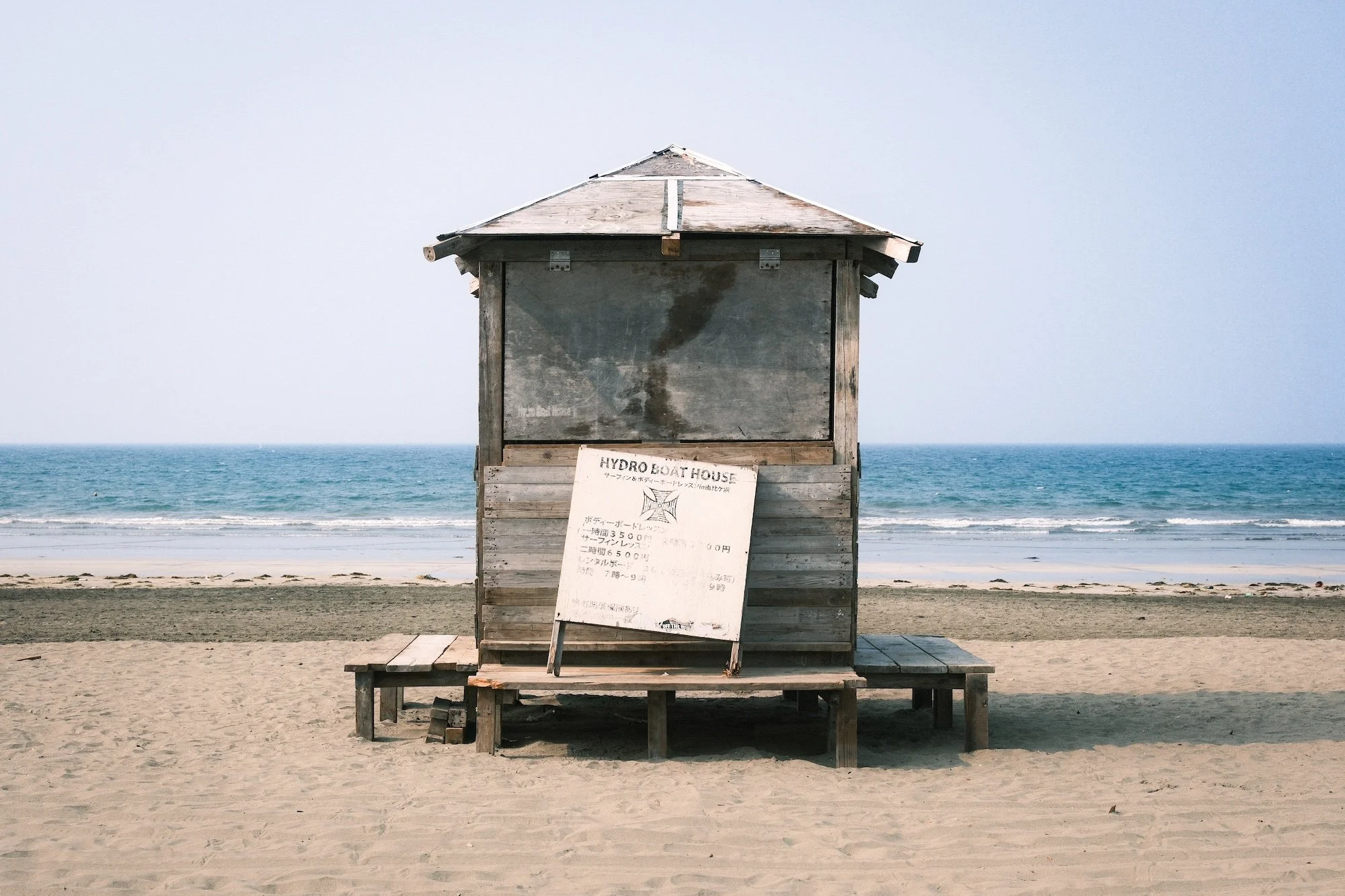 A weathered wooden lifeguard hut on a sandy beach with the ocean and blue sky in the background.