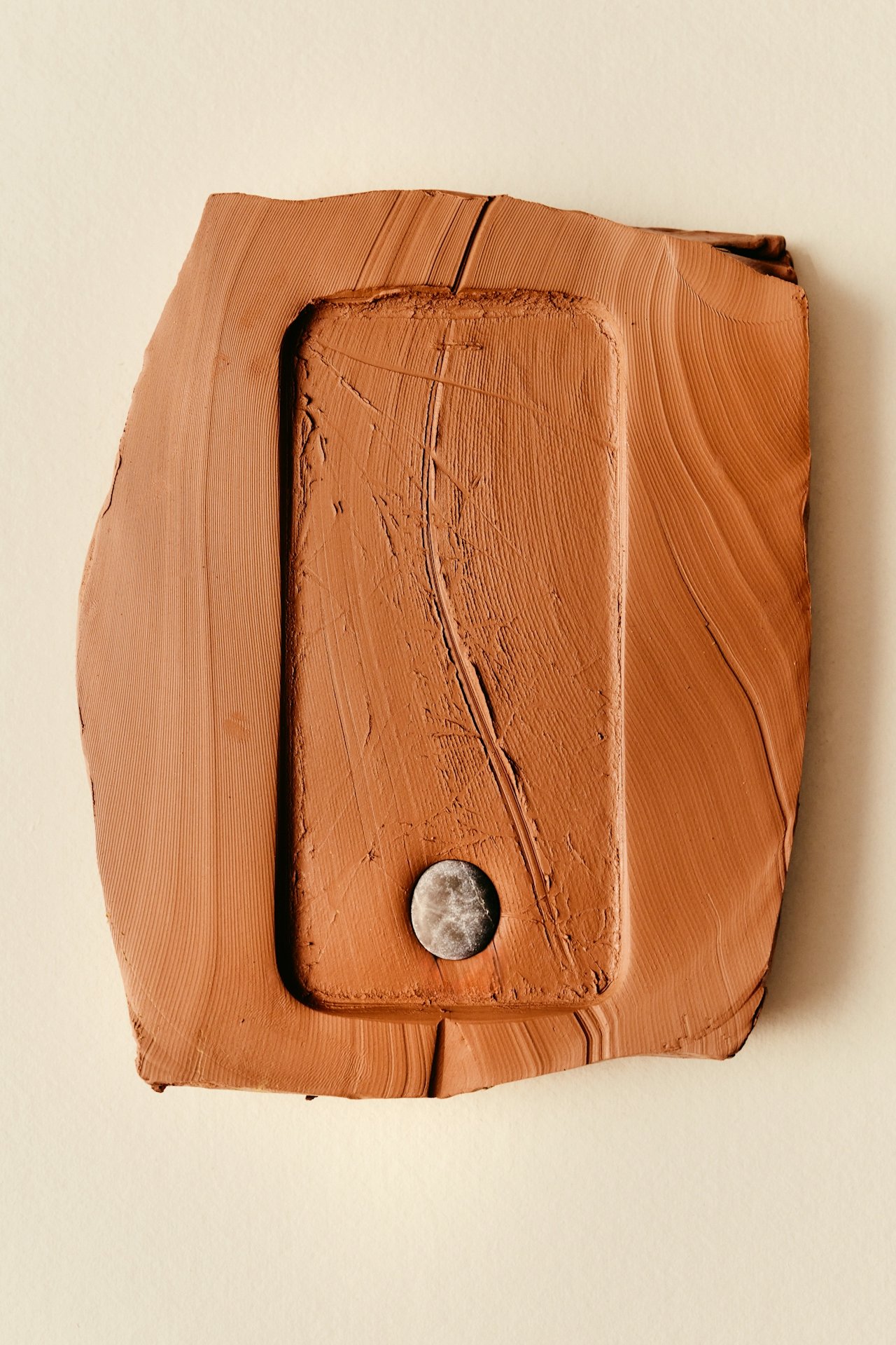 A rectangular, textured, earthy orange object made of clay or ceramic with a small round metallic element at the bottom. The object is placed against a plain light background.