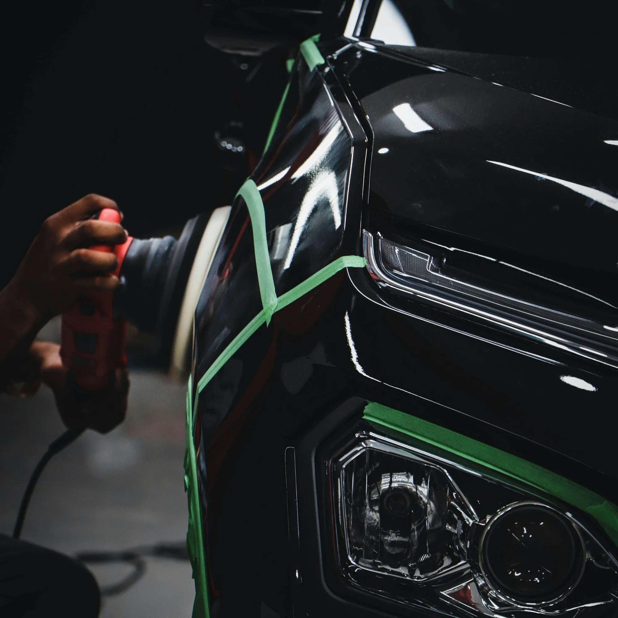 A person polishing the front of a black car with a power buffer, with green painter's tape visible on the car