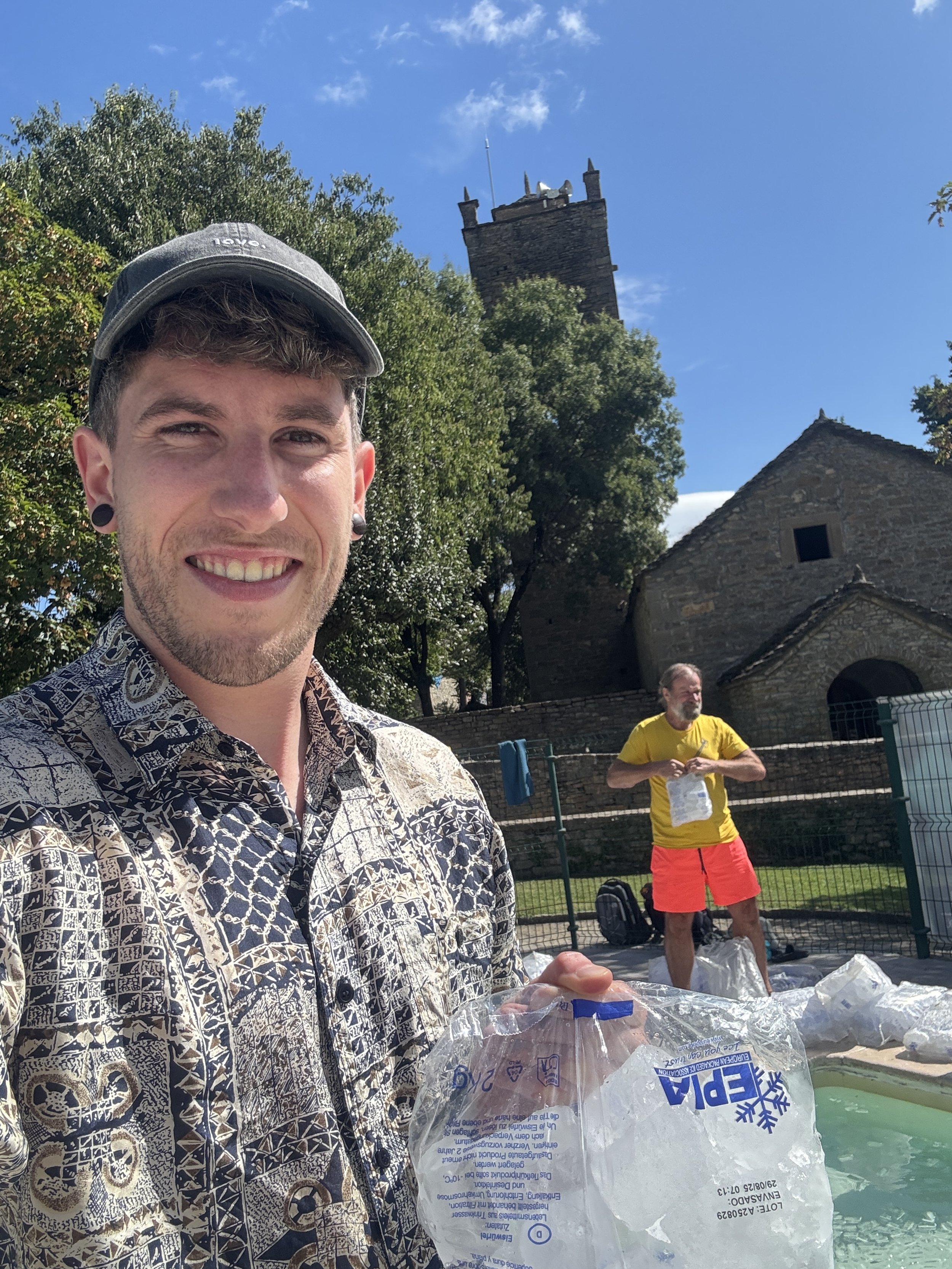 Julian Scheuermann, zertifizierter Wim Hof Method Instructor im Wim Hof Basecamp in Morillo de Tor in Spanien, hält eine Tüte Eis und posiert für das Foto, im Hintergrund steht Wim Hof und bereitet gerade das Eisbad vor.