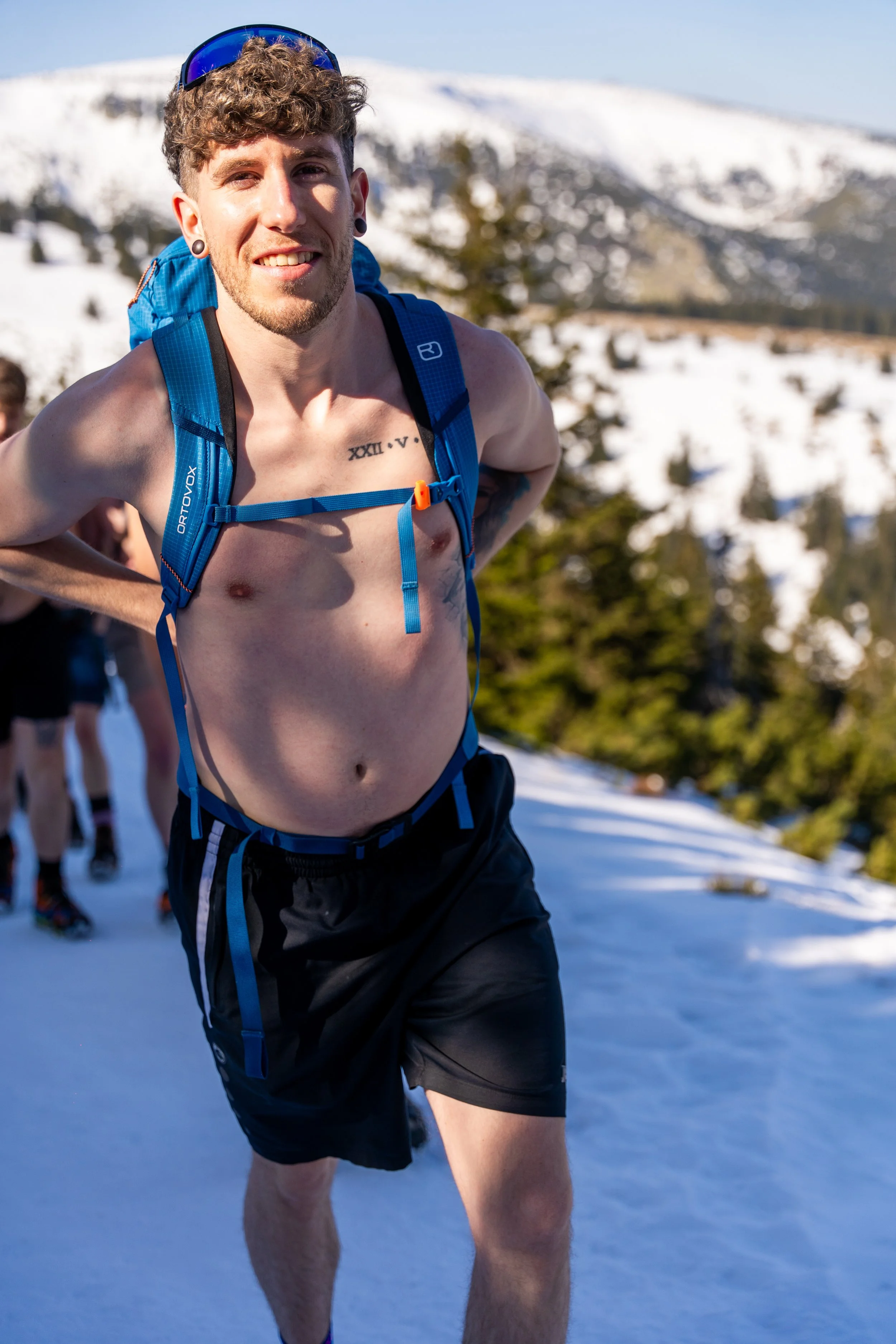Ein nackter junger Mann mit einem blauen Rucksack bei einer Kältewanderung auf Berg Śnieżka in Polen, lächelnd, mit Sonnenbrille auf dem Kopf, im Vordergrund, andere Läufer im Hintergrund.