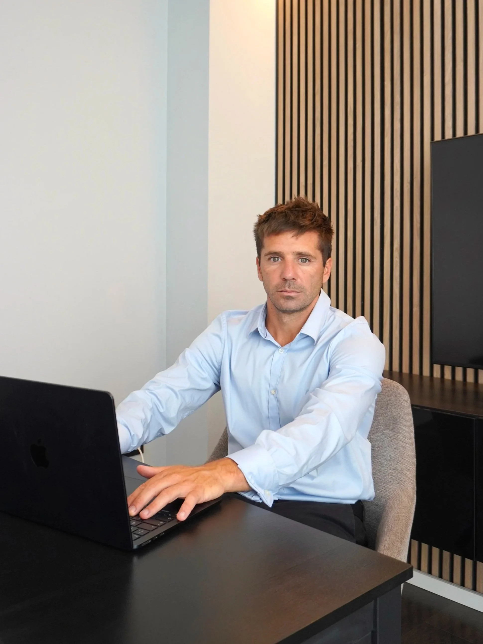 Hombre sentado en una mesa de oficina, usando una camisa azul, trabajando en una laptop, con pared de madera decorativa en el fondo.