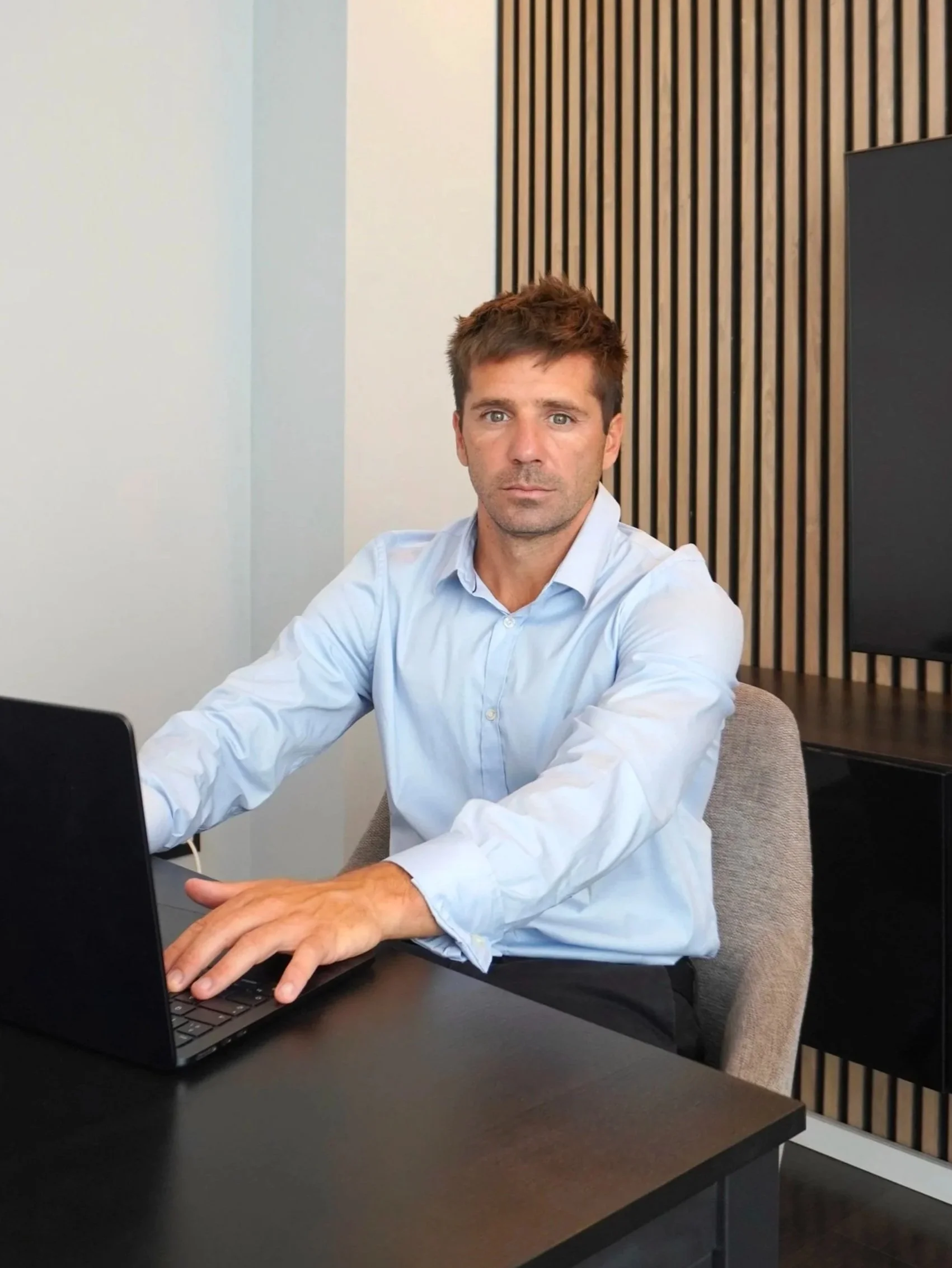 Hombre sentado en una mesa de oficina, usando una camisa azul, trabajando en una laptop, con pared de madera decorativa en el fondo.