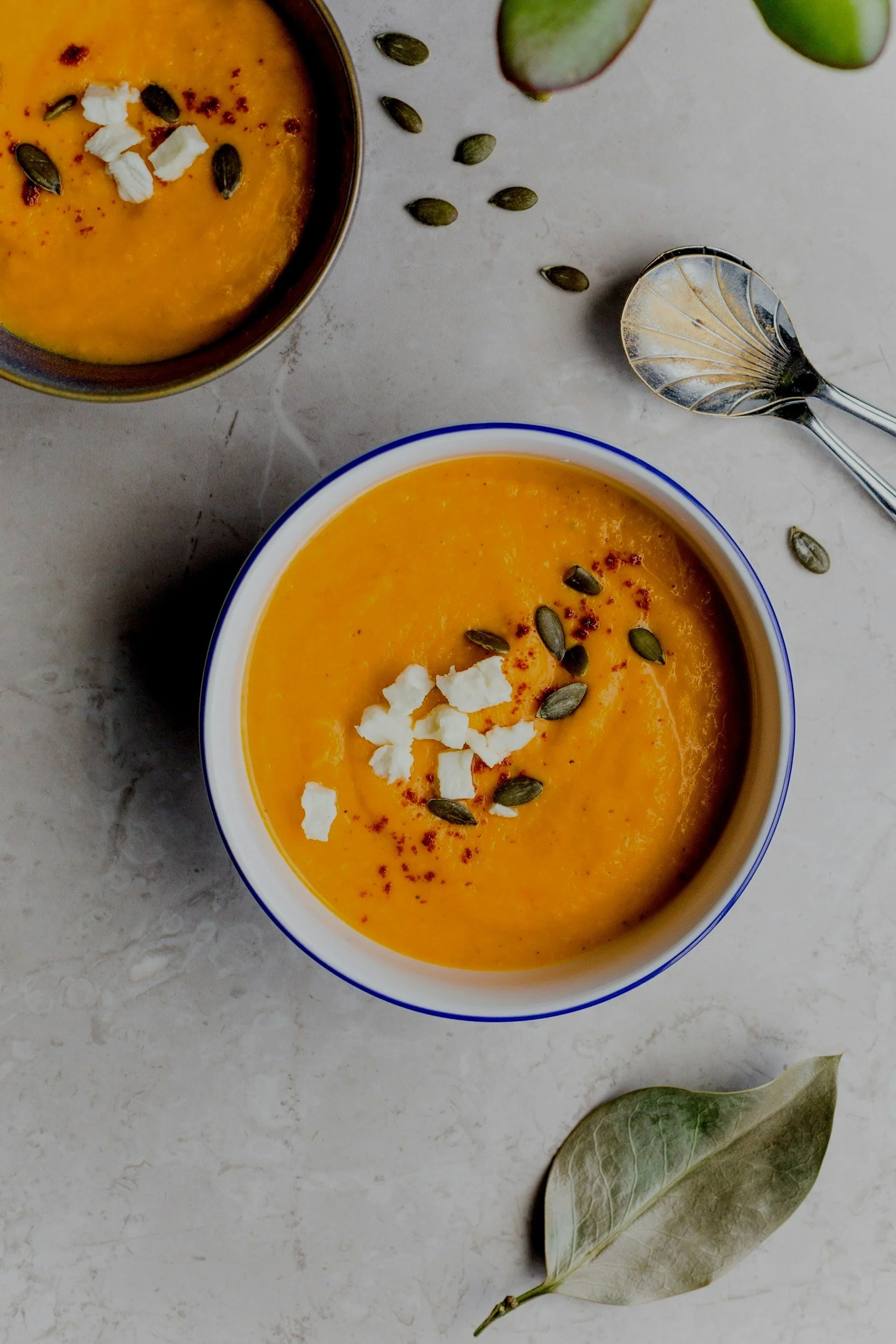 Two bowls of creamy orange soup garnished with pumpkin seeds, crumbled white cheese, and red pepper flakes, with a decorative spoon and dried leaves on a light surface.