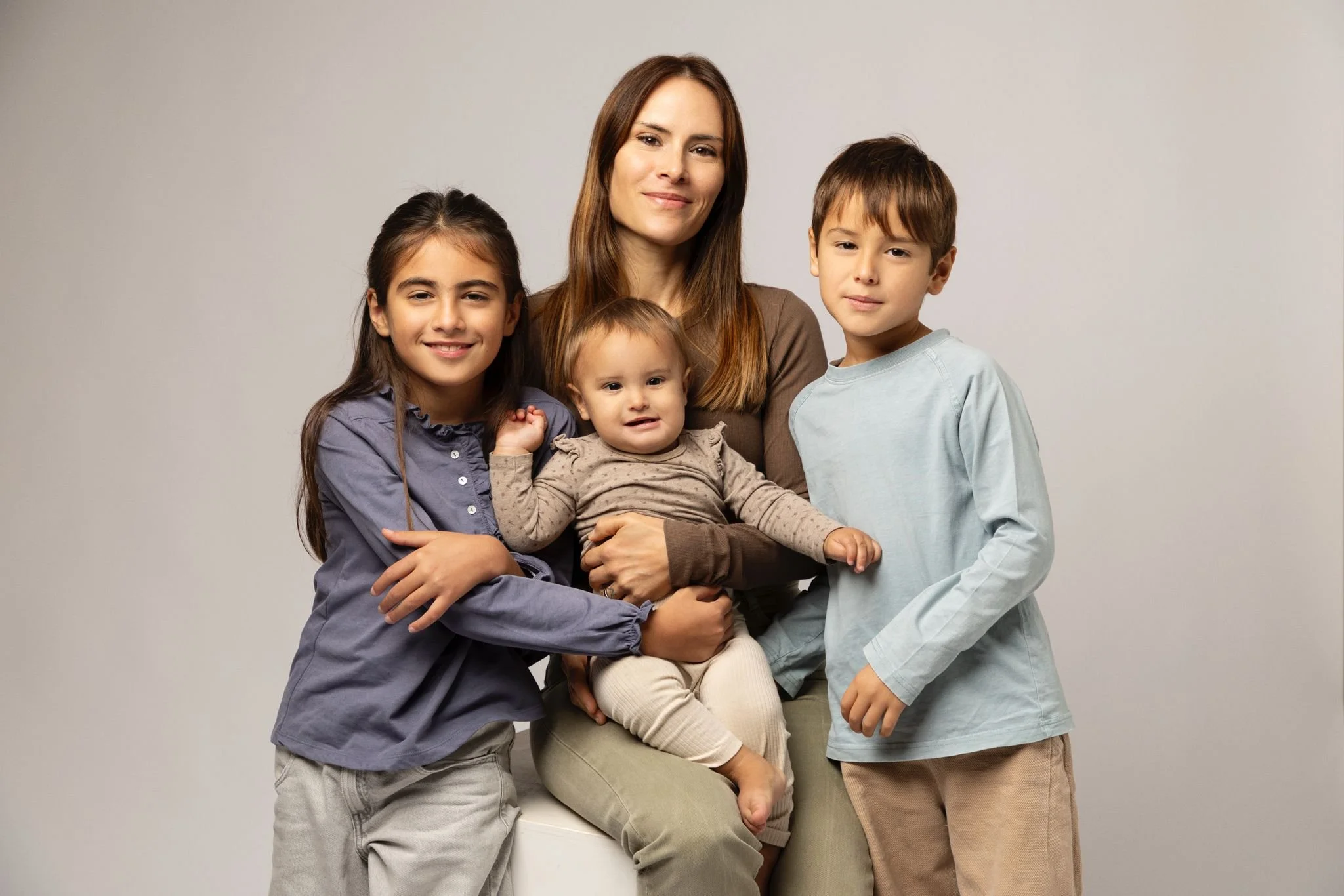 A woman with long brown hair is sitting with three children, a girl and two boys, all smiling or with neutral expressions, against a plain light-colored background.