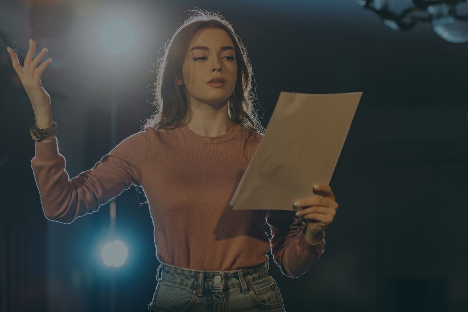 A young woman with long brown hair wearing a pink sweater and jeans, holding scripts in a theatrical setting, appears to be acting or rehearsing.