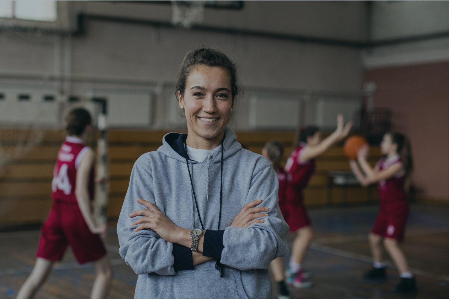 A smiling woman in a gray hoodie standing with her arms crossed in a gymnasium with girls in red basketball uniforms practicing in the background.