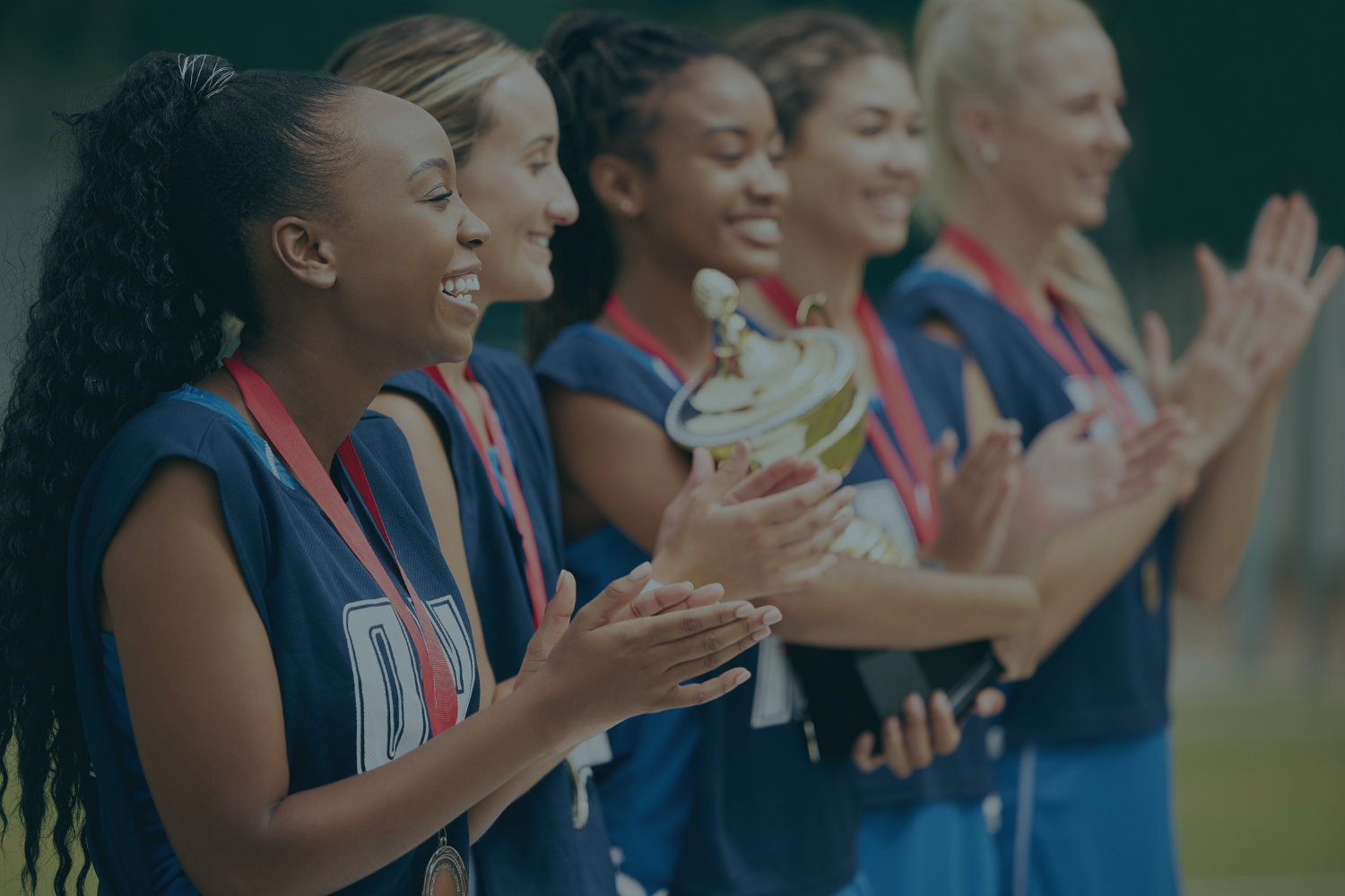A group of smiling women in sports uniforms celebrating on a sports field, with one woman holding a gold trophy.