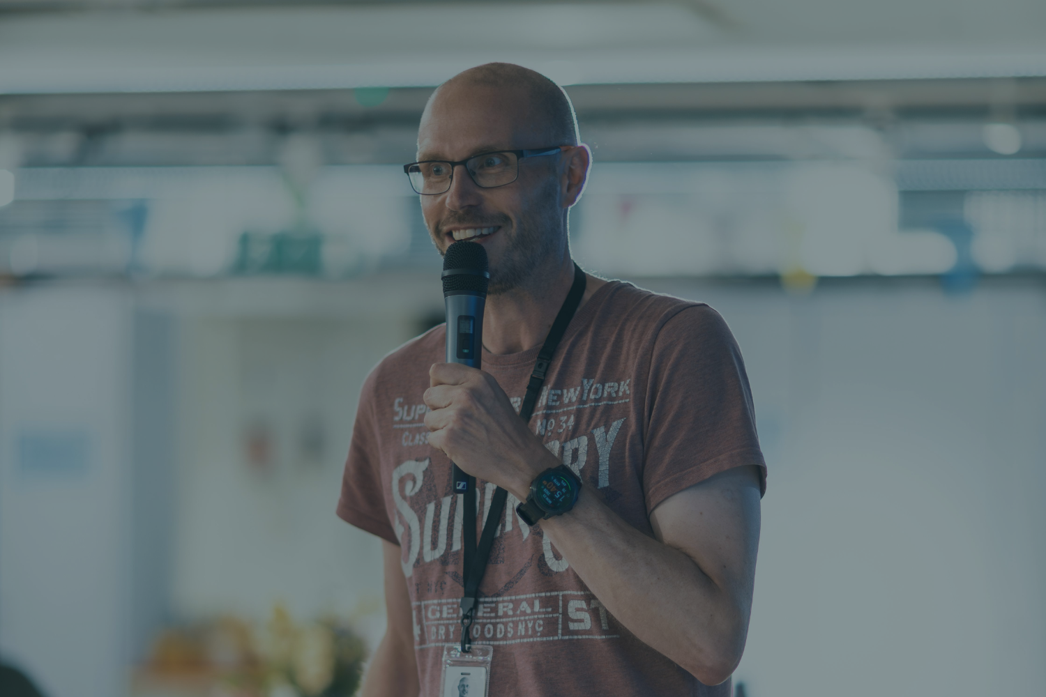 A man with glasses and a beard smiling while speaking into a microphone, wearing a watch, a lanyard, and a casual t-shirt in an indoor setting.