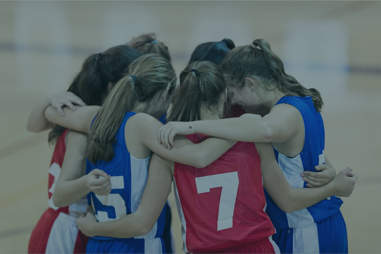 Young female basketball players in a team huddle, wearing red and blue jerseys, embracing each other in a moment of team spirit on the court.