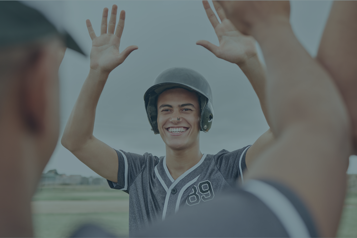 A young man in a baseball jersey and a helmet smiling and raising his hands for a high five with two other people outdoors.