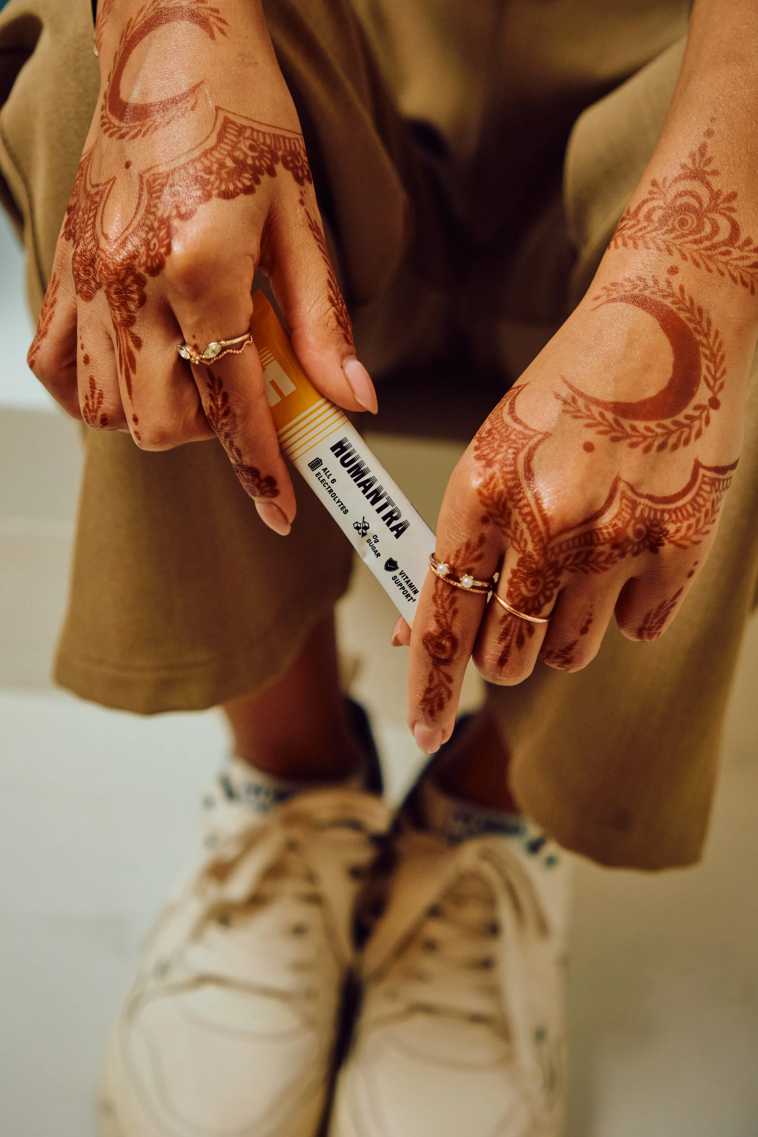 Person with henna tattoos on hands, wearing rings, holding a container labeled "Humanika" dietary supplement, wearing tan pants and white sneakers.