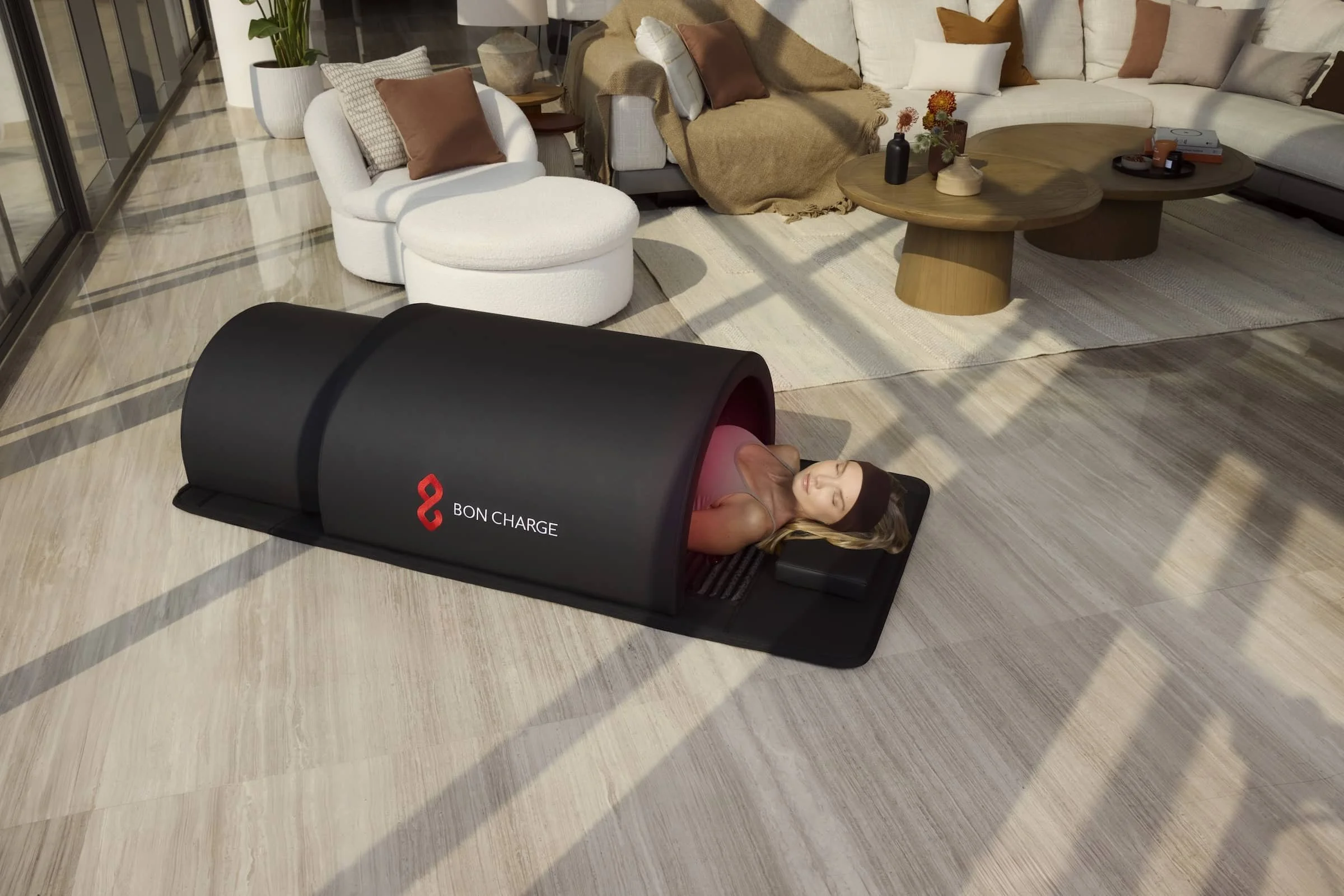 A woman lying inside a black portable sauna tent labeled 'Bon Charge' in a living room, with sunlight shining through large windows.