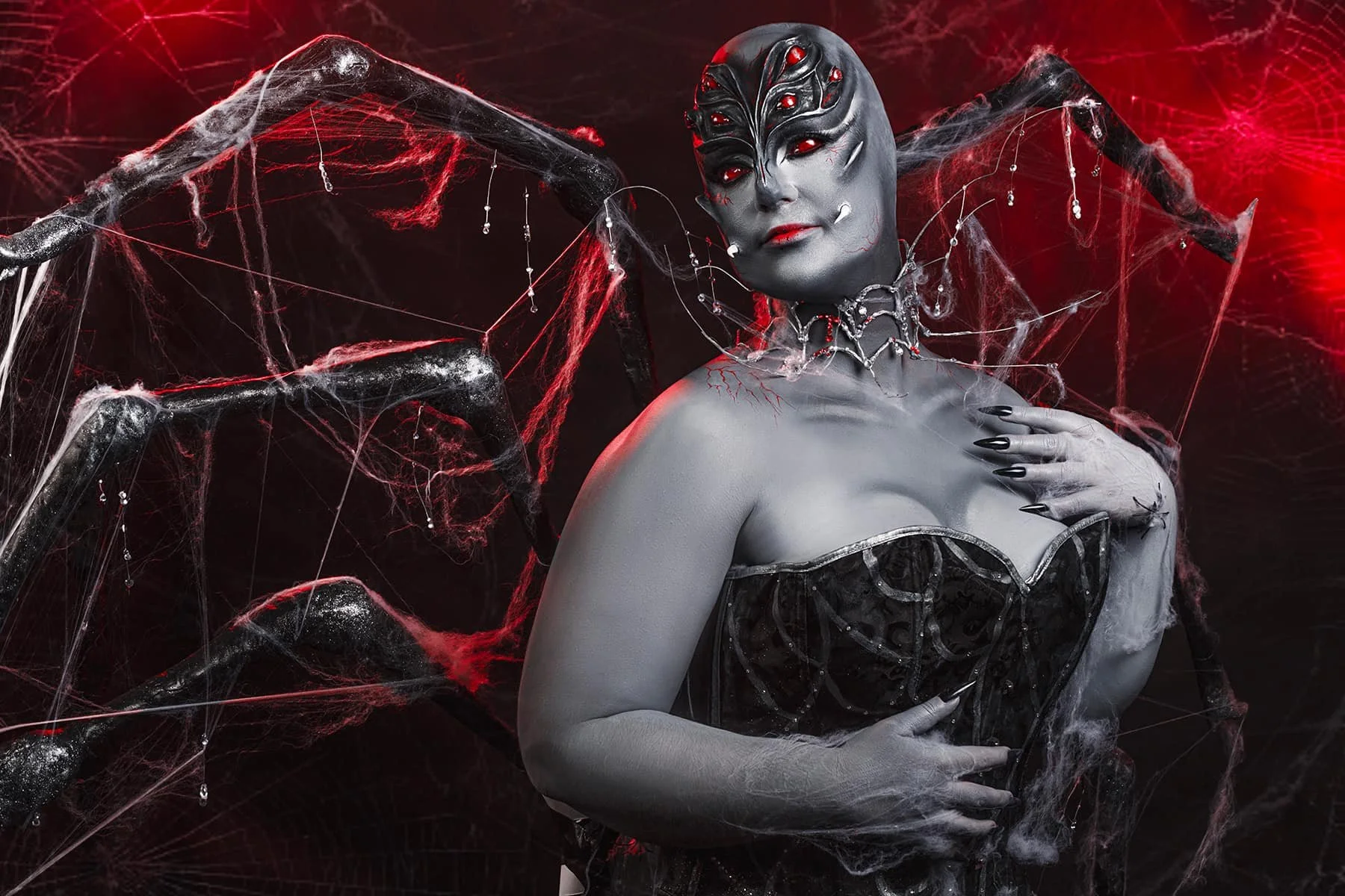 A woman in dark, gothic makeup and costume with spider motifs, surrounded by spider webs and black spiders with red eyes, in a dark red and black background.