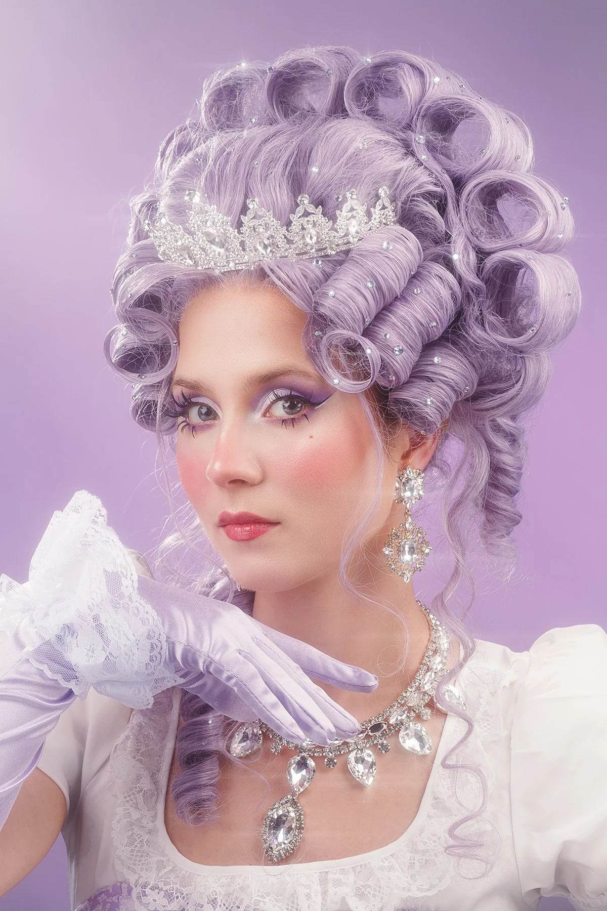 A woman with light purple curly hair styled in an 18th-century fashion, adorned with a silver tiara. She is wearing dramatic purple eye makeup, pearl accents in her hair, large crystal earrings, a matching necklace, and a white lace dress with a puffed sleeve. She is also wearing a white lace glove on her left hand, which is resting near her face, against a purple background.