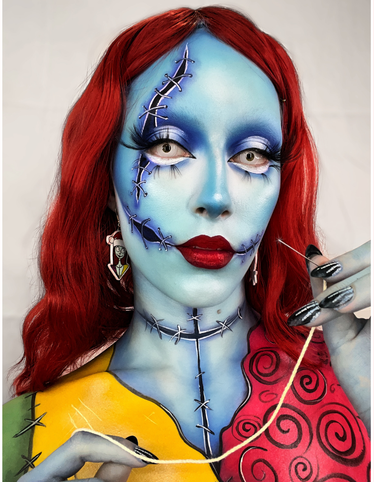 A woman with elaborate makeup resembling Sally from The Nightmare Before Christmas, featuring blue face paint, stitched smile design, and striking red hair, holding a needle and thread.
