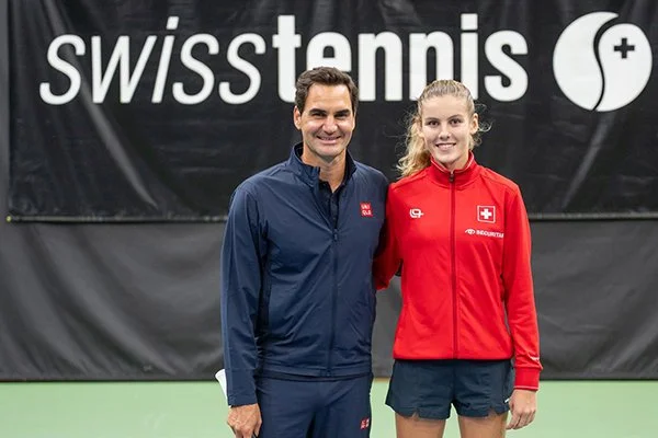 Ein Mann und eine junge Frau stehen nebeneinander vor einem schwarzen Hintergrund mit dem Schriftzug 'swisstennis'. Der Mann trägt eine dunkelblaue Jacke, die Frau trägt eine rote Jacke mit Schweizer Flagge und schwarzen Shorts.