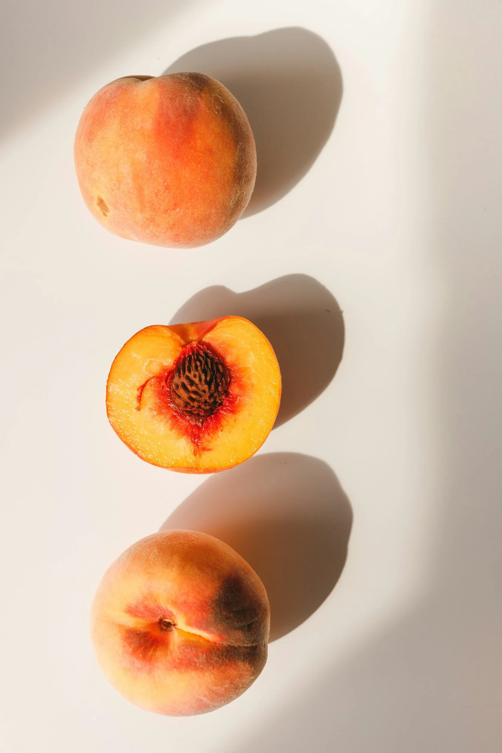 Three peaches, one whole at the top, one cut in half revealing the pit and flesh, and one whole at the bottom, on a white surface with shadows.