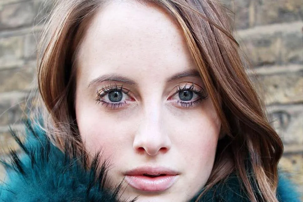 Rosie Fortescue wearing Boudoir Lashes by Asma Docrat A close-up portrait of a young woman with red hair, blue eyes, and fair skin, wearing a teal coat with a black fur collar, standing outdoors in front of a brick wall.