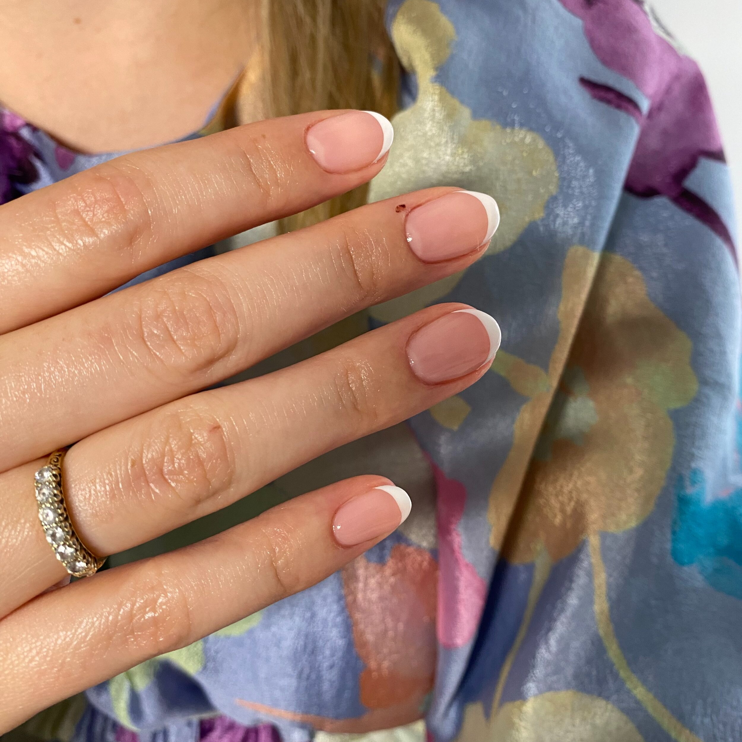 Close-up of a hand with a diamond wedding ring, showing freshly manicured nails with French tips, against a colorful floral fabric background.