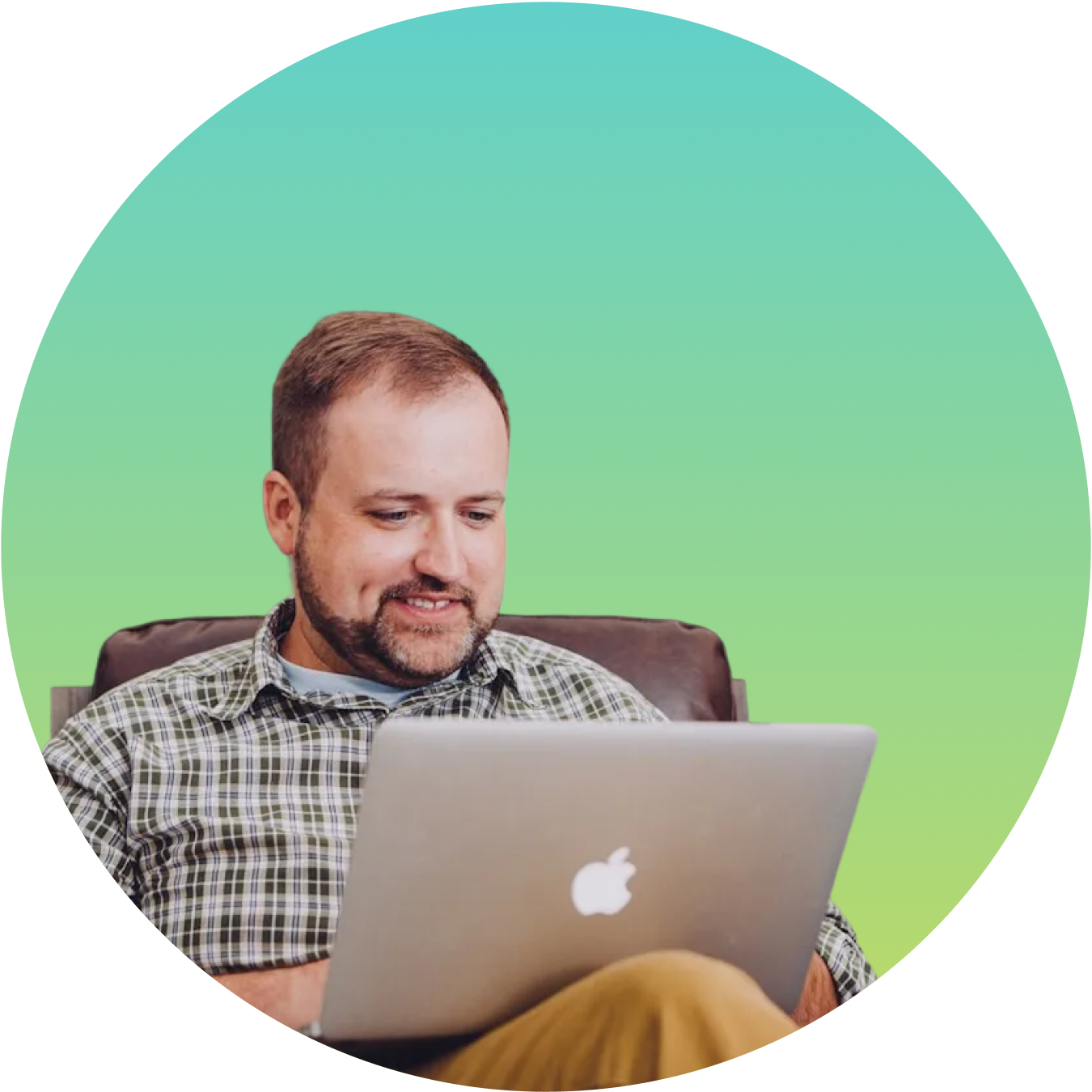 A man with short brown hair and a beard, wearing a plaid shirt, sitting on a brown chair and smiling while looking at his open MacBook laptop.