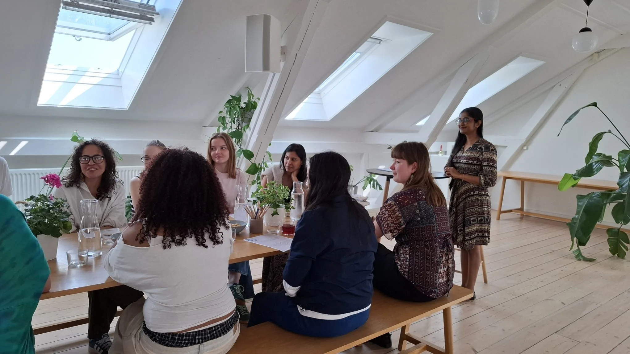 Group of women sitting around a wooden table in a bright room with skylights, plants, and a standing woman speaking.