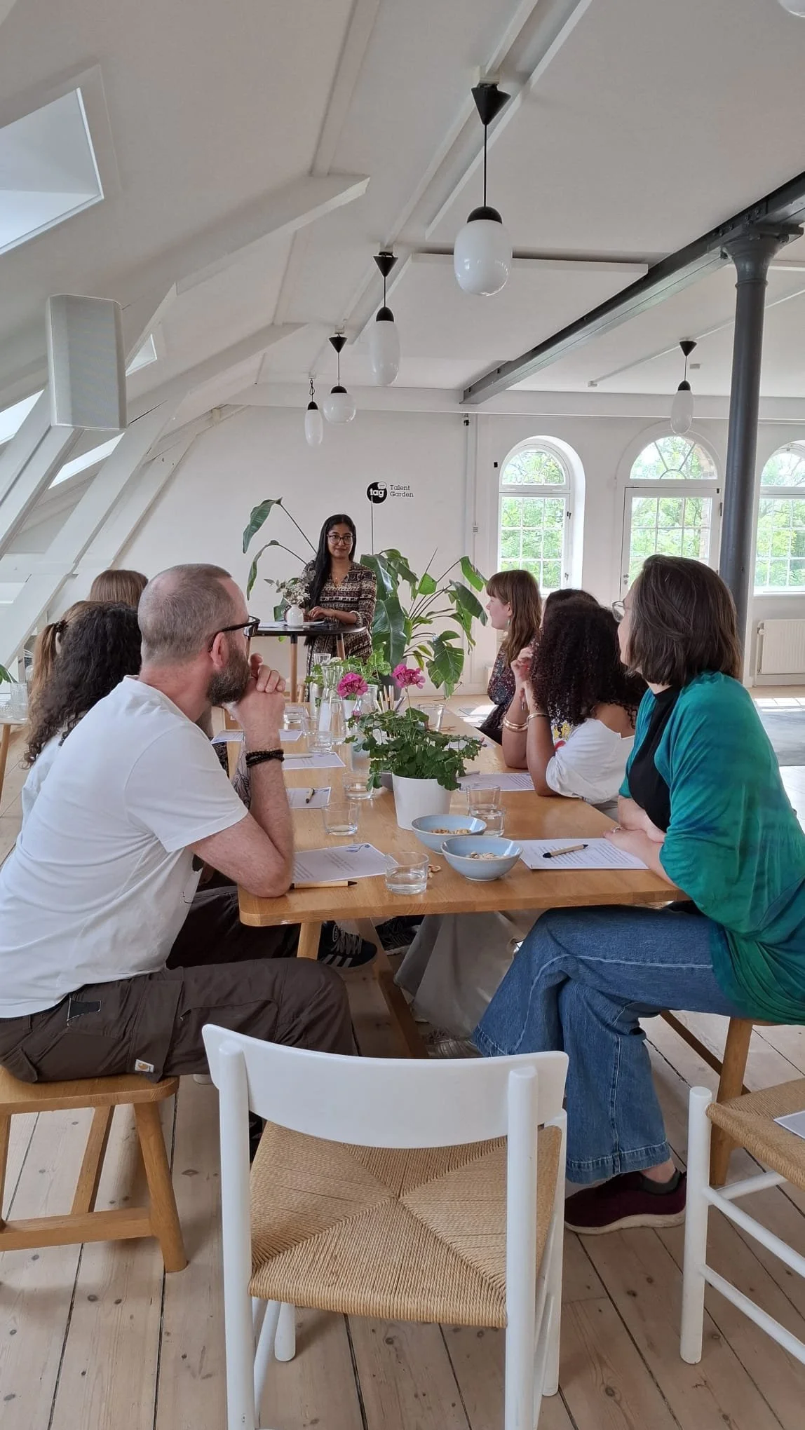 A group of people sitting around a wooden table, attentive to a woman standing and speaking at the front of the room. The setting is a bright room with large windows, hanging lights, and potted plants.
