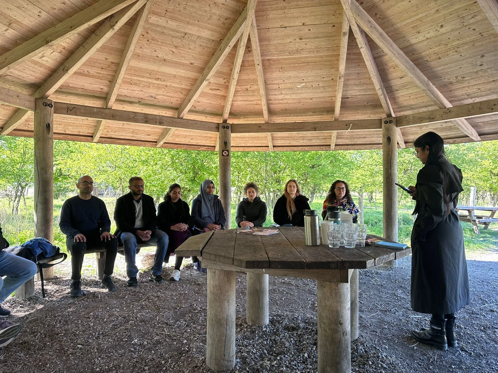 Group of seven people sitting in a semi-circle under a wooden gazebo, listening to a woman standing at a table with drinks and materials, outdoors with green trees in the background.