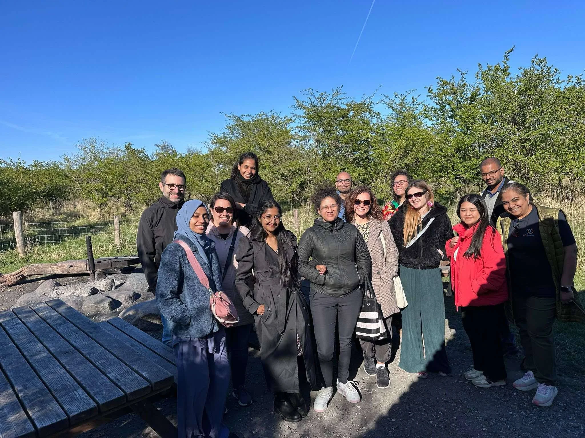 Group of diverse people outdoors on a sunny day, standing on a trail with trees and bushes in the background, smiling and posing for a photo.