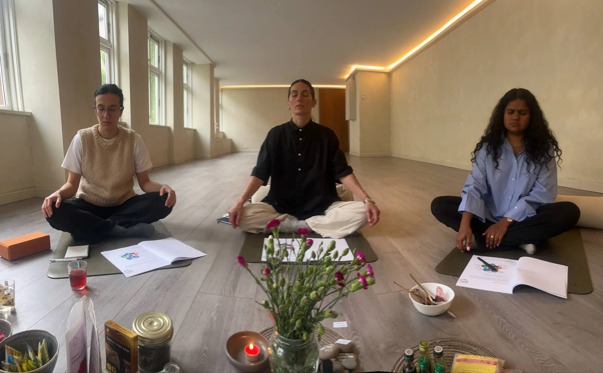 Three women sit cross-legged on yoga mats in a spacious room with large windows, meditating with closed eyes, surrounded by books, flowers, and various items on the floor.