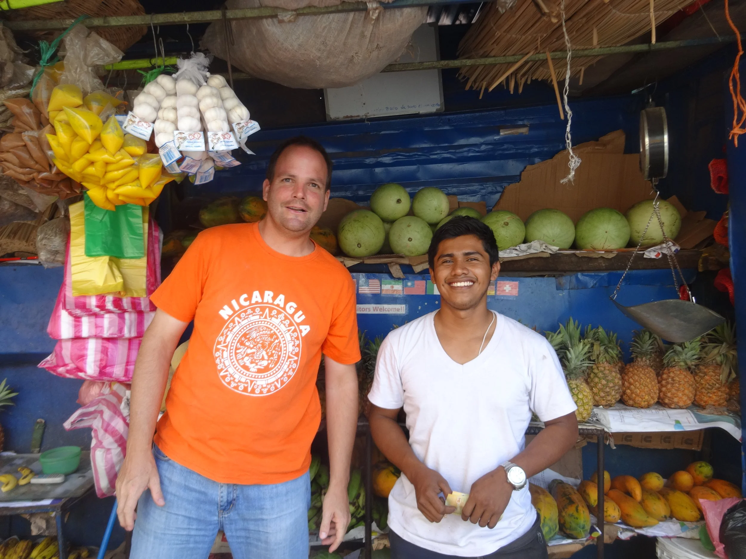 Ehemaliger Langzeitmitarbeitender im orangen T-Shirt steht mit einem lokalen Früchteverkäufer vor dessen Verkaufsstand. Stay-Mitarbeitende berichten von ihren Erfahrungen während ihres Langzeiteinsatzes in Lateinamerika.