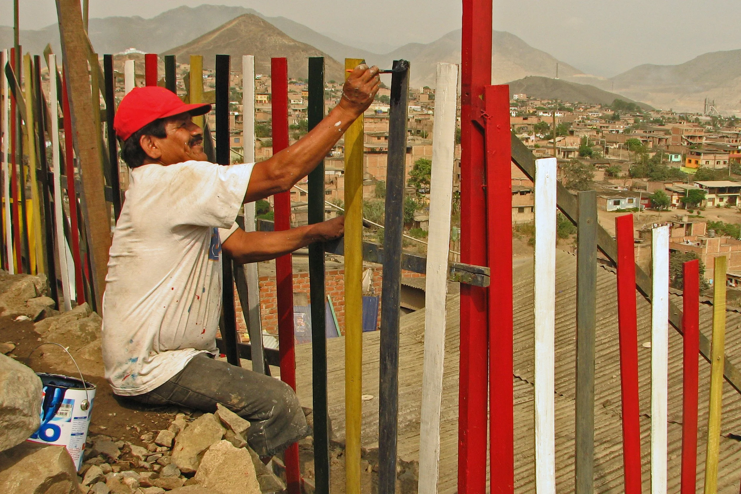Ein Mann streicht einen Zaun in leuchtenden Farben – im Hintergrund die trockene Landschaft von Lima, Peru. Symbol für Stay-Einsätze bei Latin Link: Langzeiteinsätze nach einem 2-jährigen Kurzzeiteinsatz.