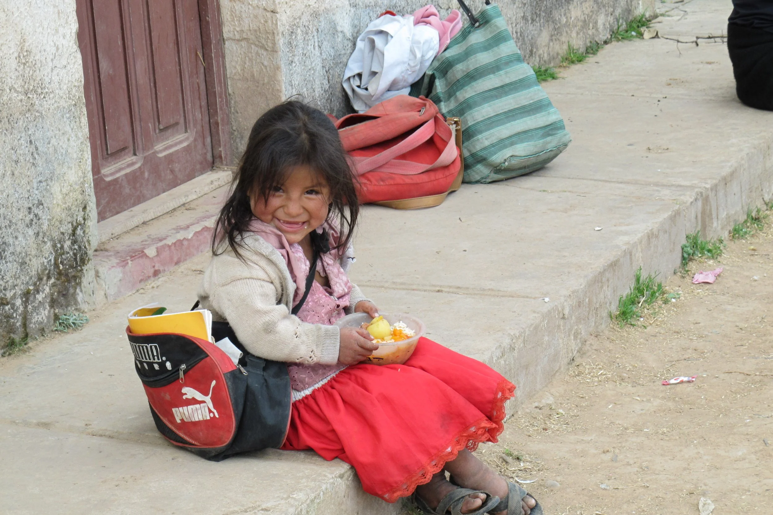 Lächelndes kleines Mädchen mit Essenschüssel auf dem Schoss – zeigt Lebensrealitäten in Südamerika, wie sie Mitarbeitende von Latin Link während Team-, Kurz- oder Langzeiteinsätzen erleben.